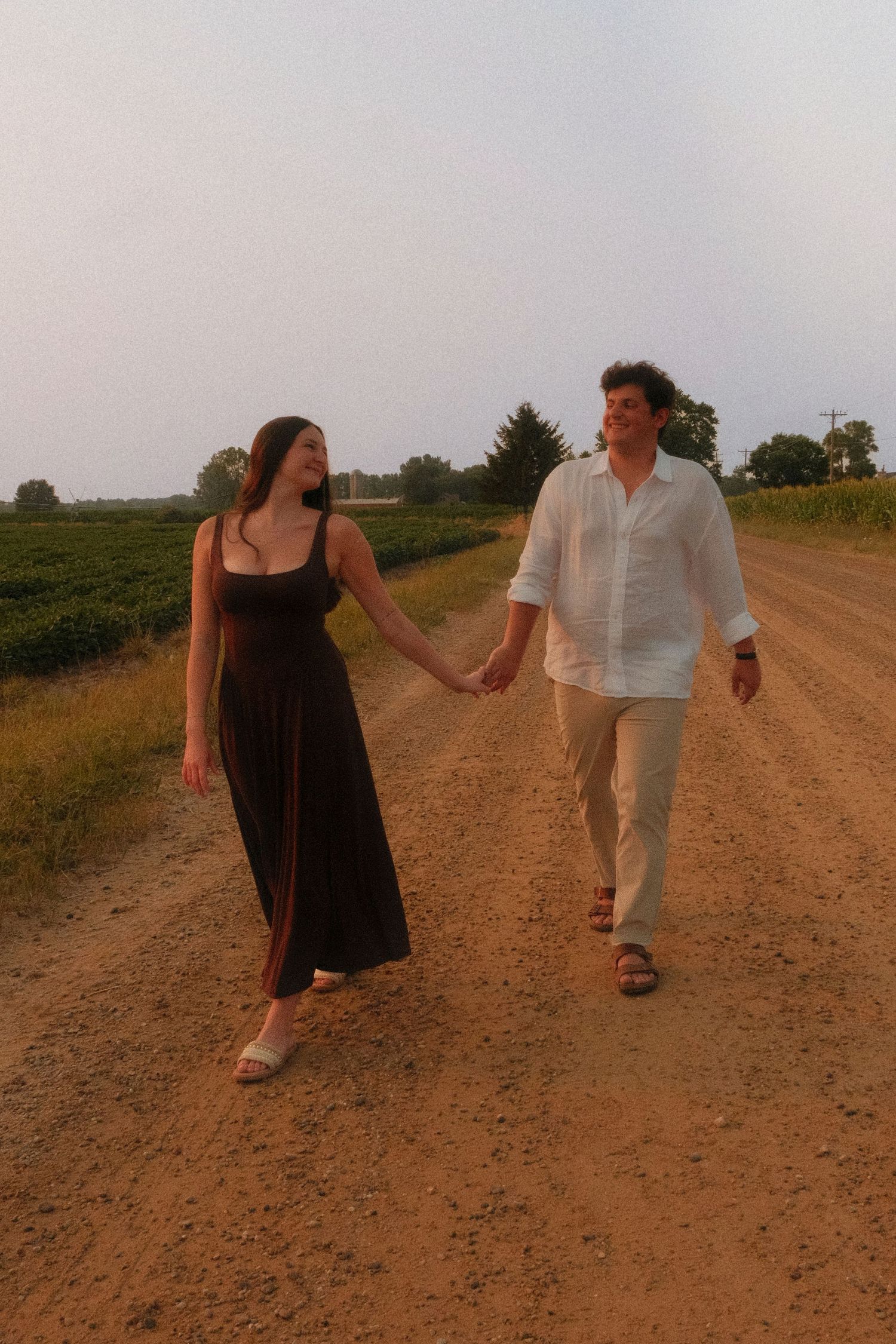 A couple holds hands walking down a rural dirt road at sunset, woman in black maxi dress, man in white linen shirt.