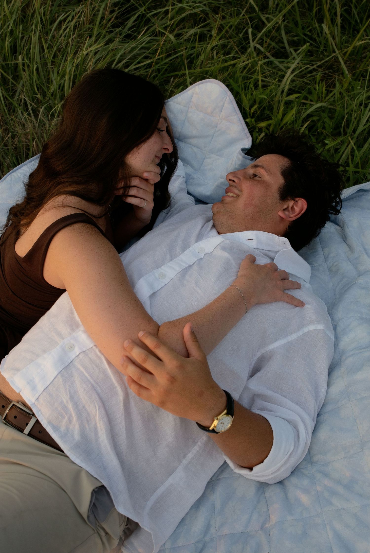 Couple lying on a blanket in green grass, smiling and embracing each other affectionately.