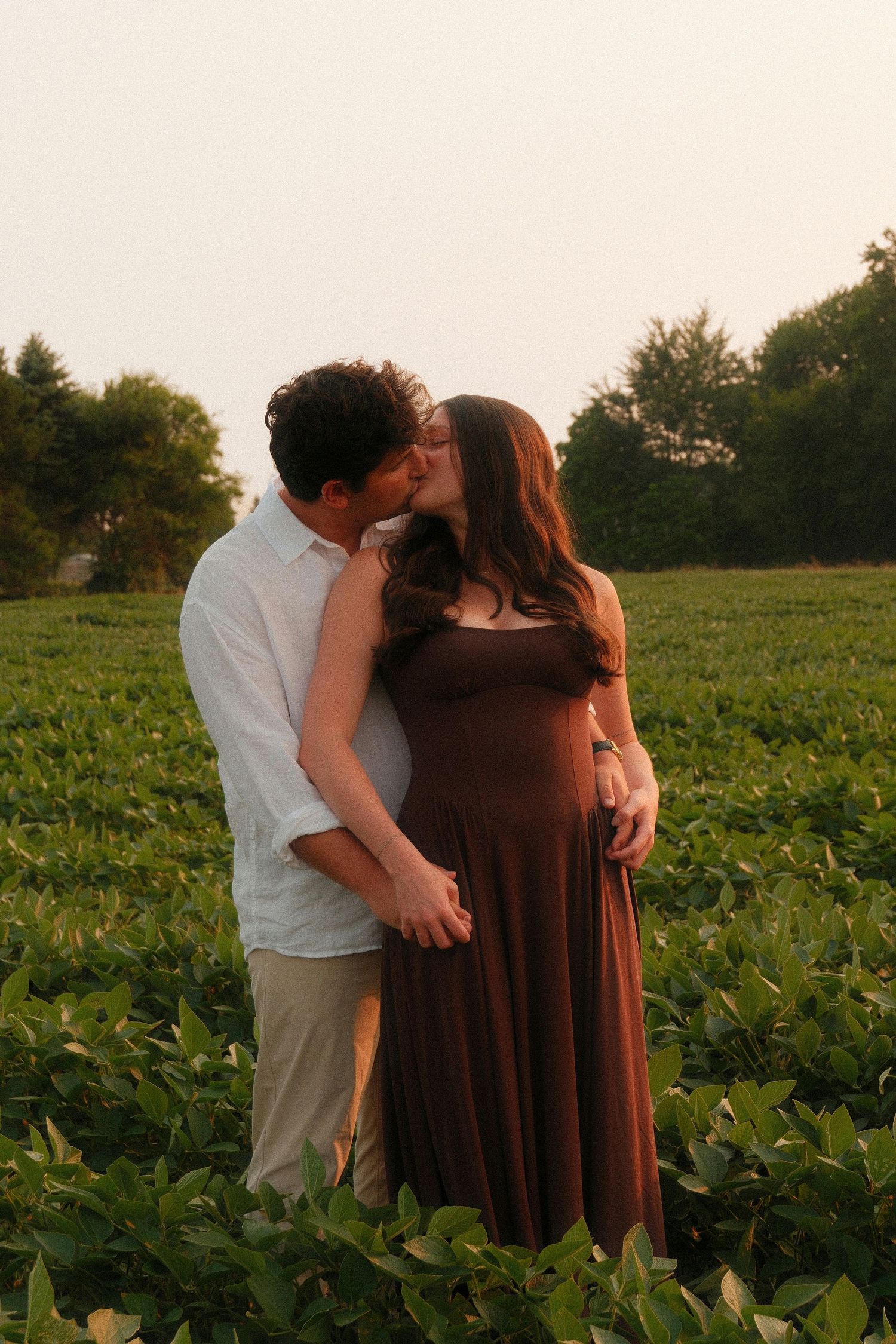 Pregnant couple sharing a romantic kiss in a lush green field at golden hour.