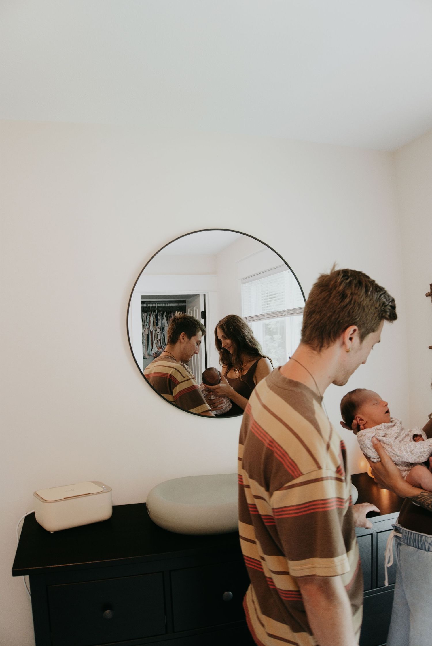 A man in a striped shirt holds a newborn baby, with a couple's reflection visible in a round mirror on a white wall.