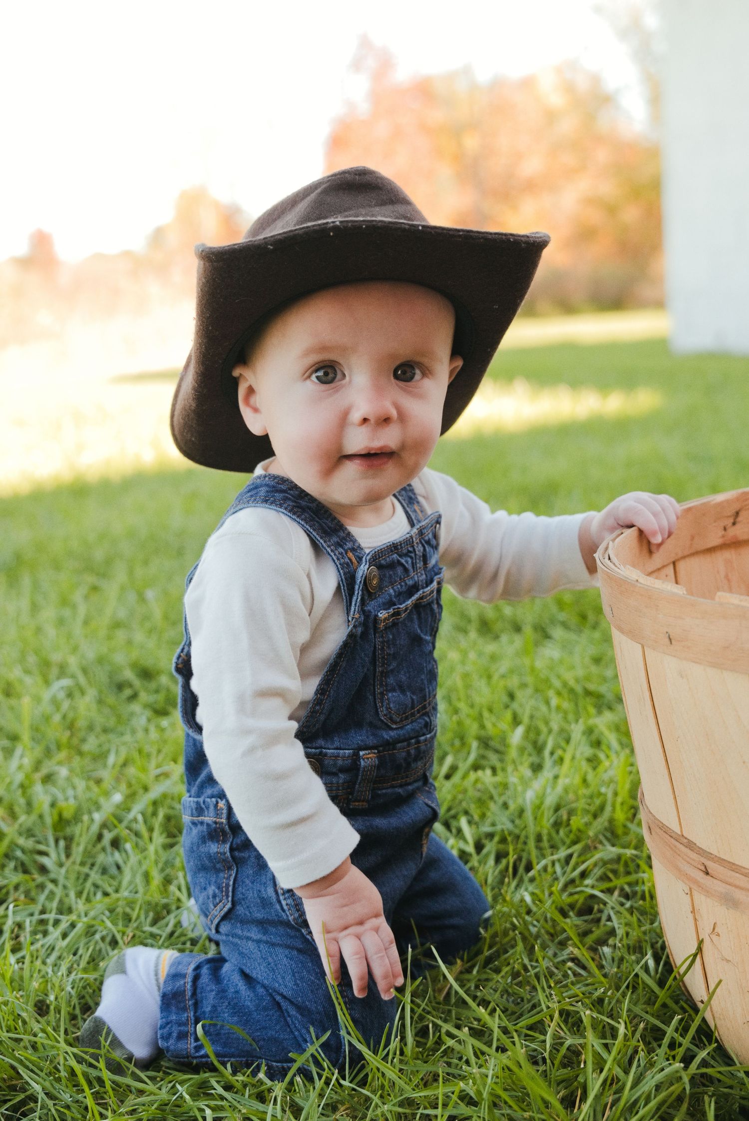 Baby in a brown cowboy hat and denim overalls kneeling on green grass beside a wooden apple basket on a sunny autumn day.