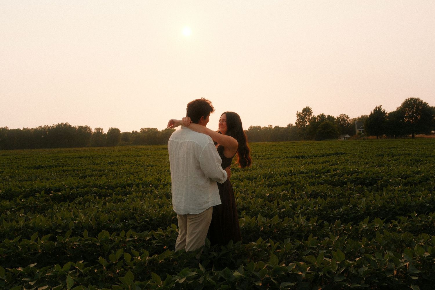 Couple embracing romantically in a lush green field at sunset with trees in the background.