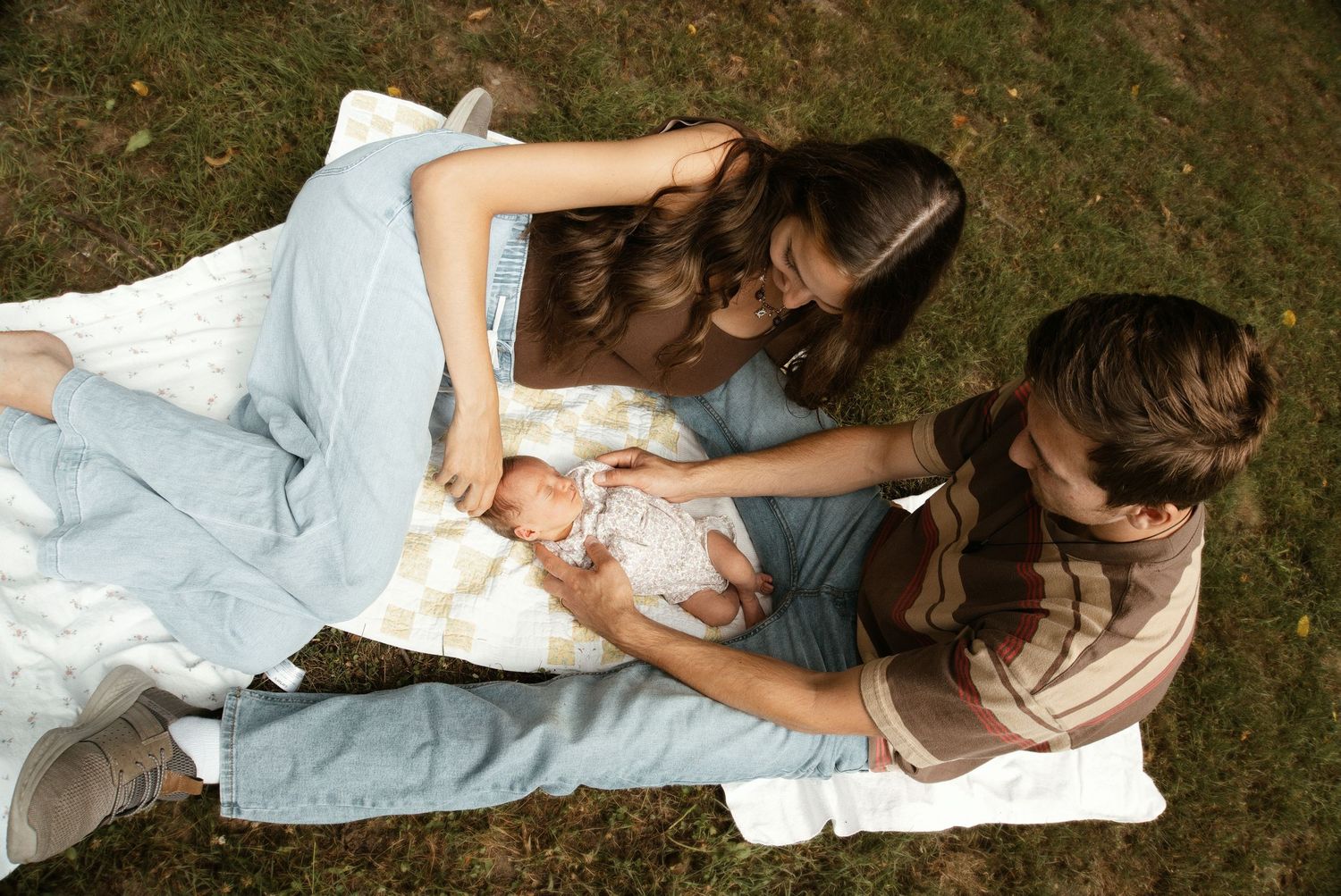Two young women sit on a blanket in grass, lovingly tending to a newborn baby outdoors.