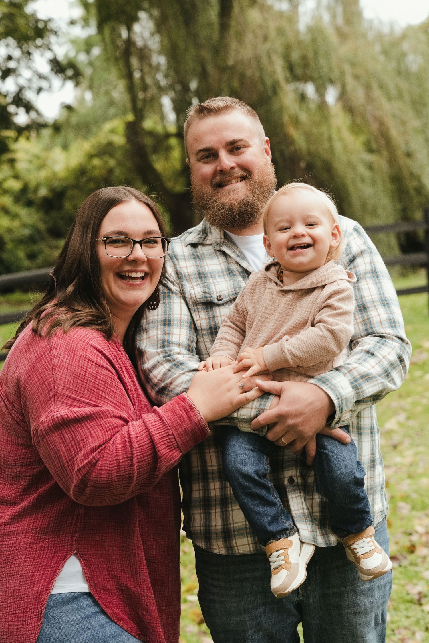 Smiling bearded man in a plaid shirt and woman with glasses in a red sweater hold a laughing baby outdoors in a green park.