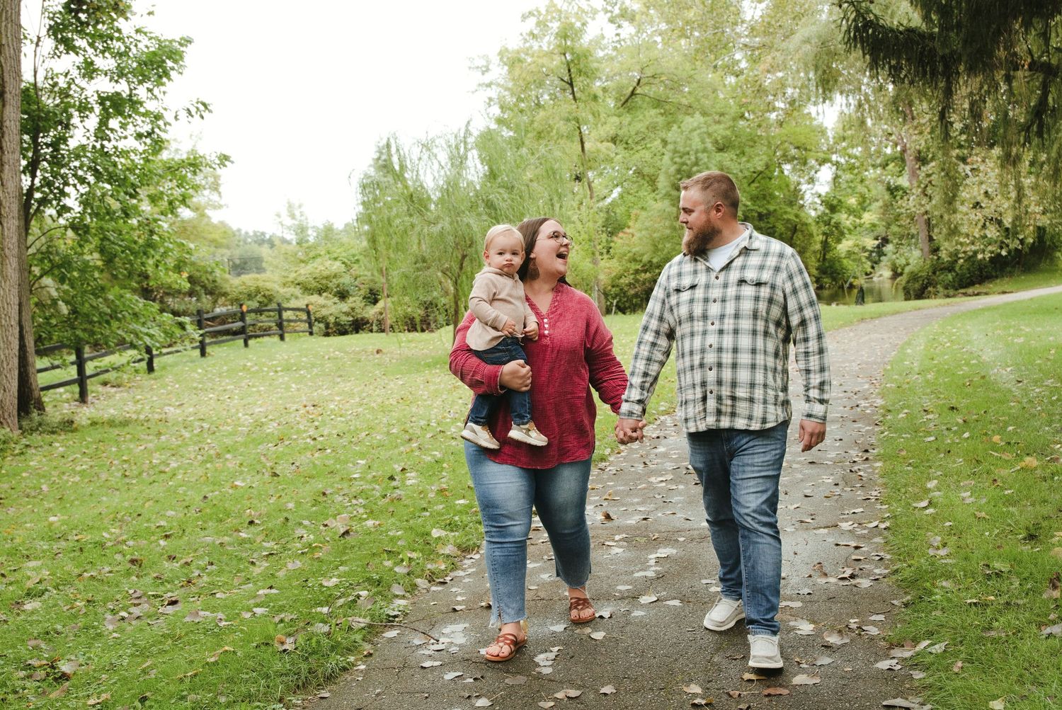 Smiling couple holding hands and walking along a leaf-covered park path while the woman carries a toddler, surrounded by green trees.