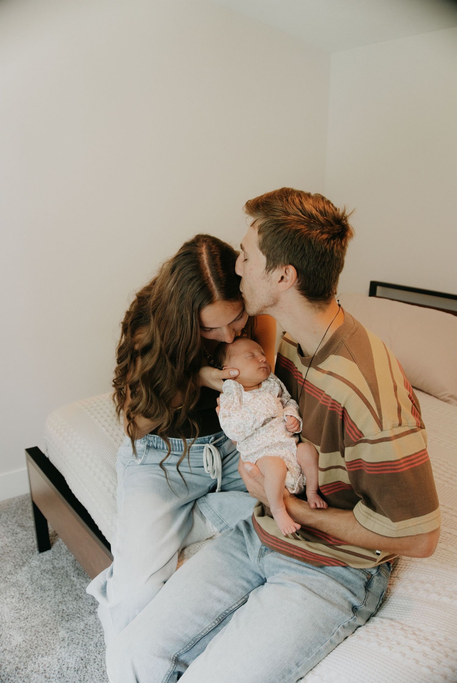 Young couple sharing a kiss over their newborn baby while sitting together on a bed.