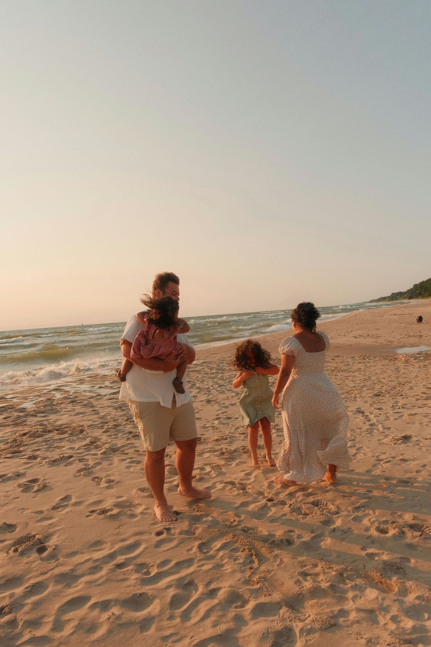 Family of four walking barefoot on sandy beach at sunset, parents with two children enjoying ocean waves.