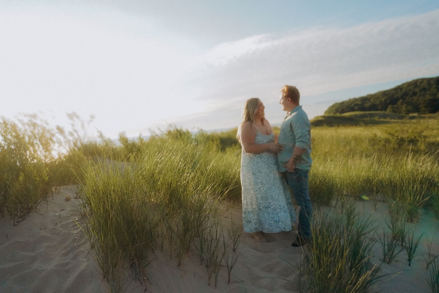 Couple standing together in sandy dunes with tall green grass at golden hour near a serene coastal landscape.