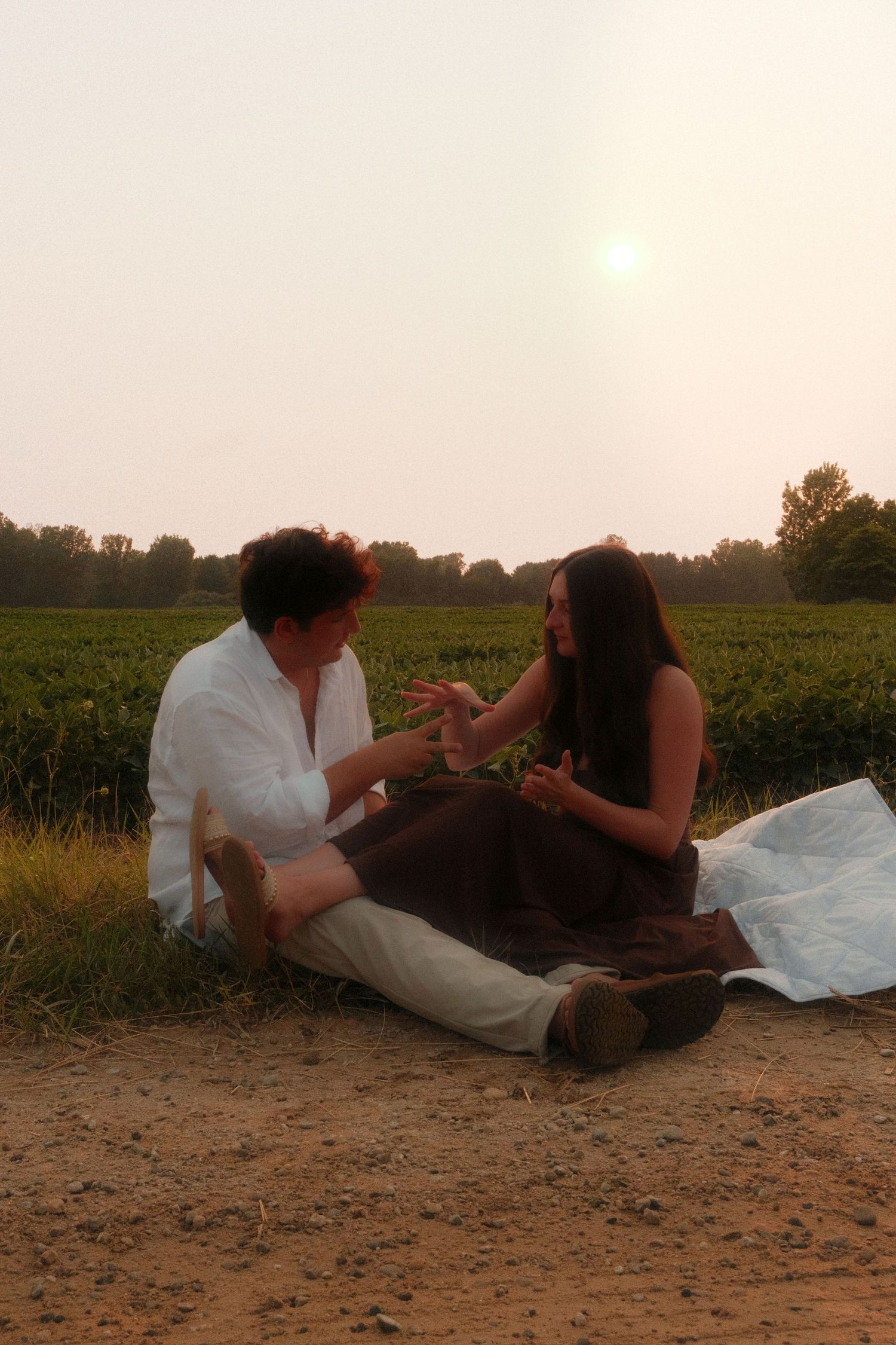 Couple sitting on a dirt path beside lush green fields, engaged in an intimate conversation at sunset.