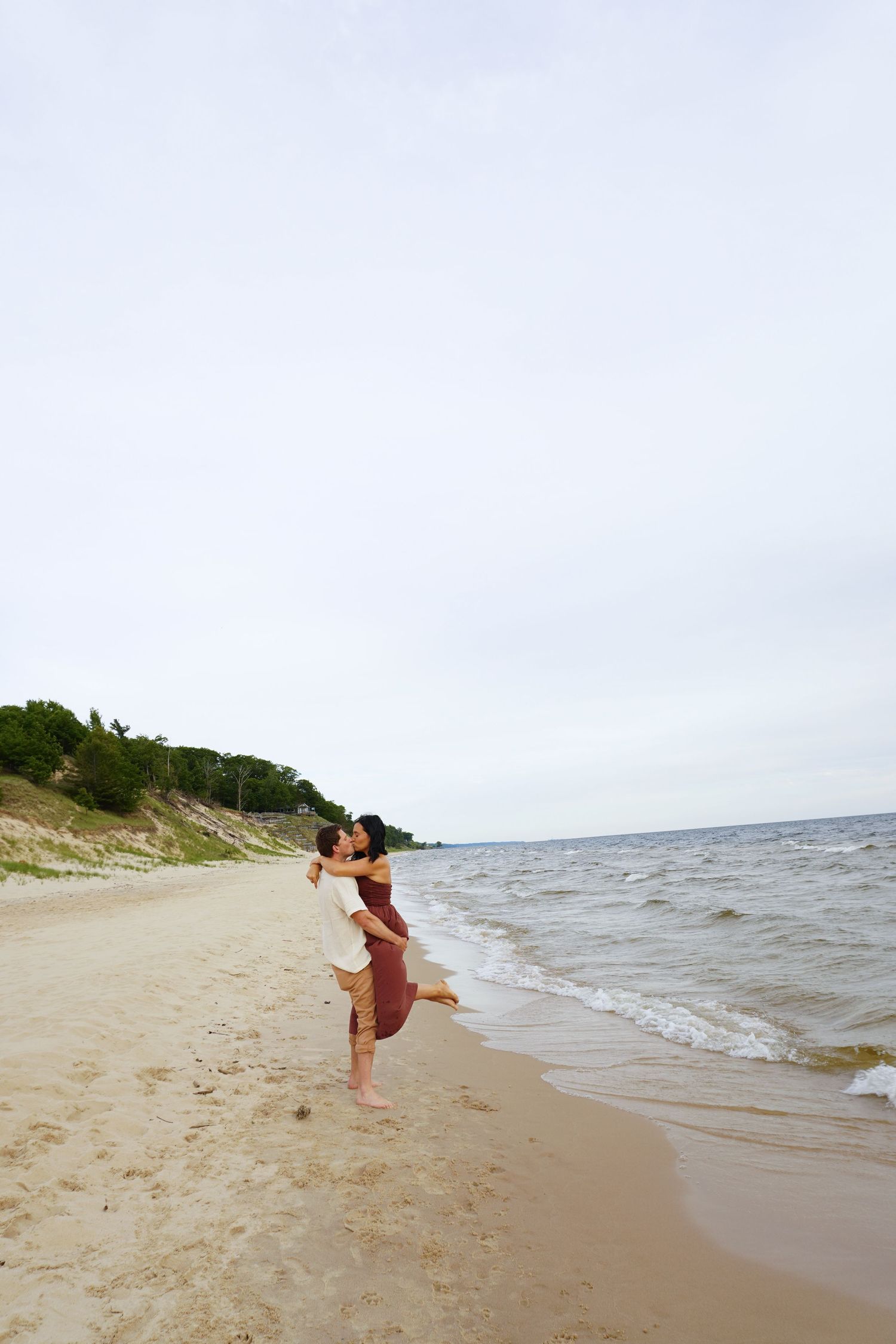 Couple embracing and spinning on a sandy beach shoreline with gentle waves and cloudy sky in background.