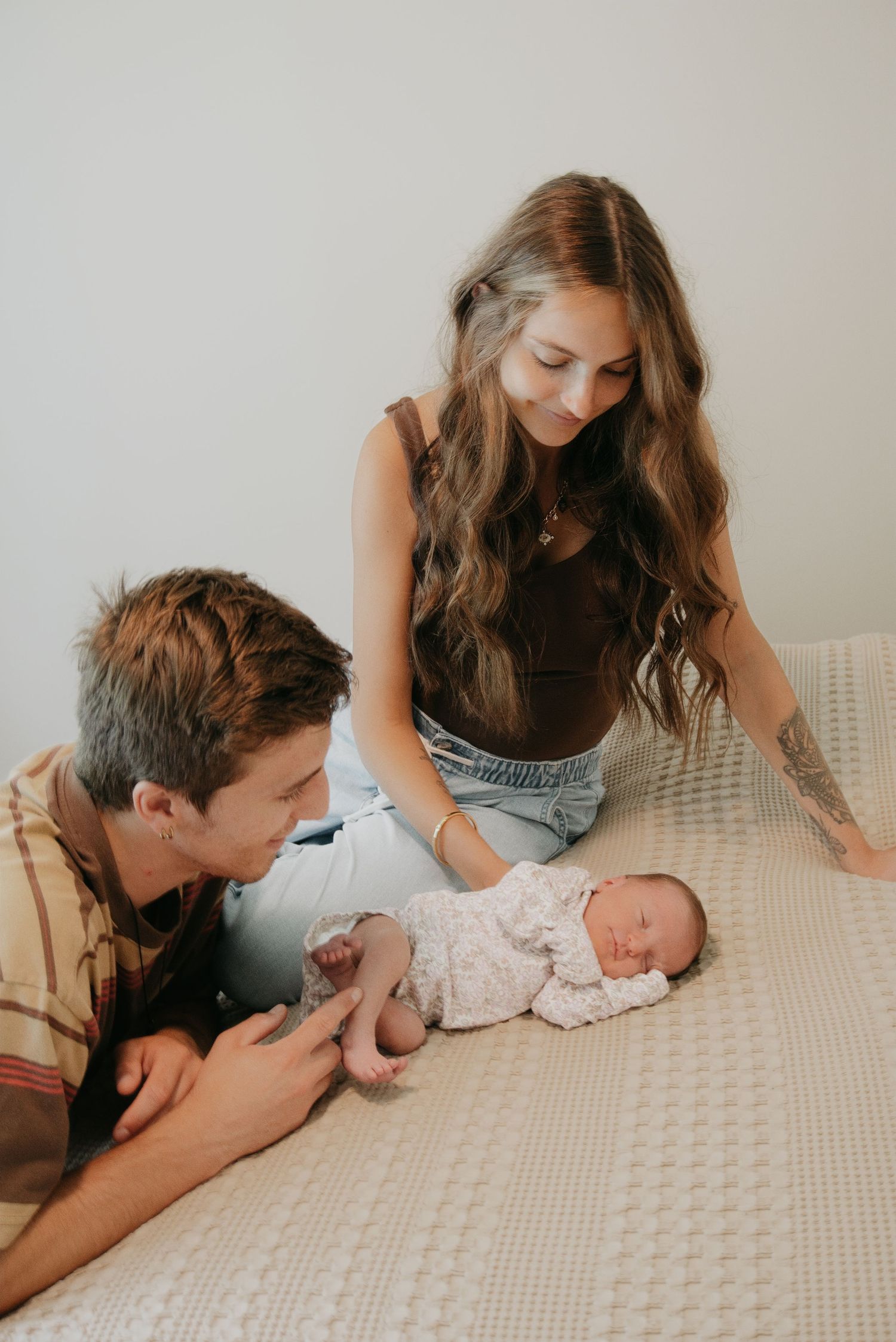 Young parents lovingly gaze at their sleeping newborn baby lying on a white knit blanket on a bed.