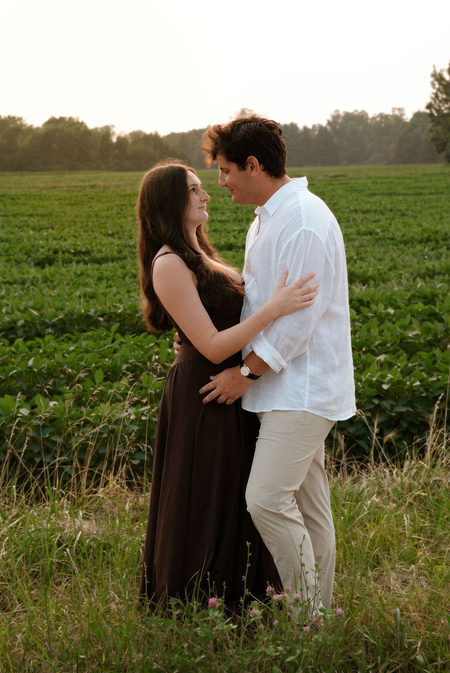 Romantic couple holding each other in a green field at golden hour, woman in brown dress and man in white shirt.