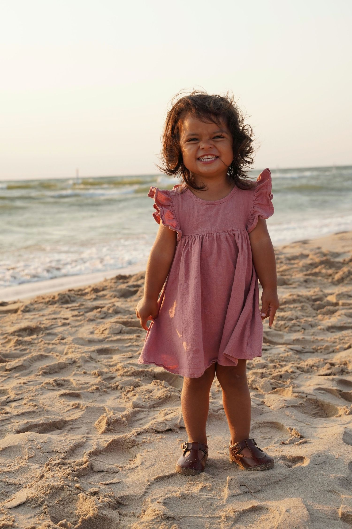 Cute toddler girl in pink ruffle dress standing on sandy beach with ocean waves in background.