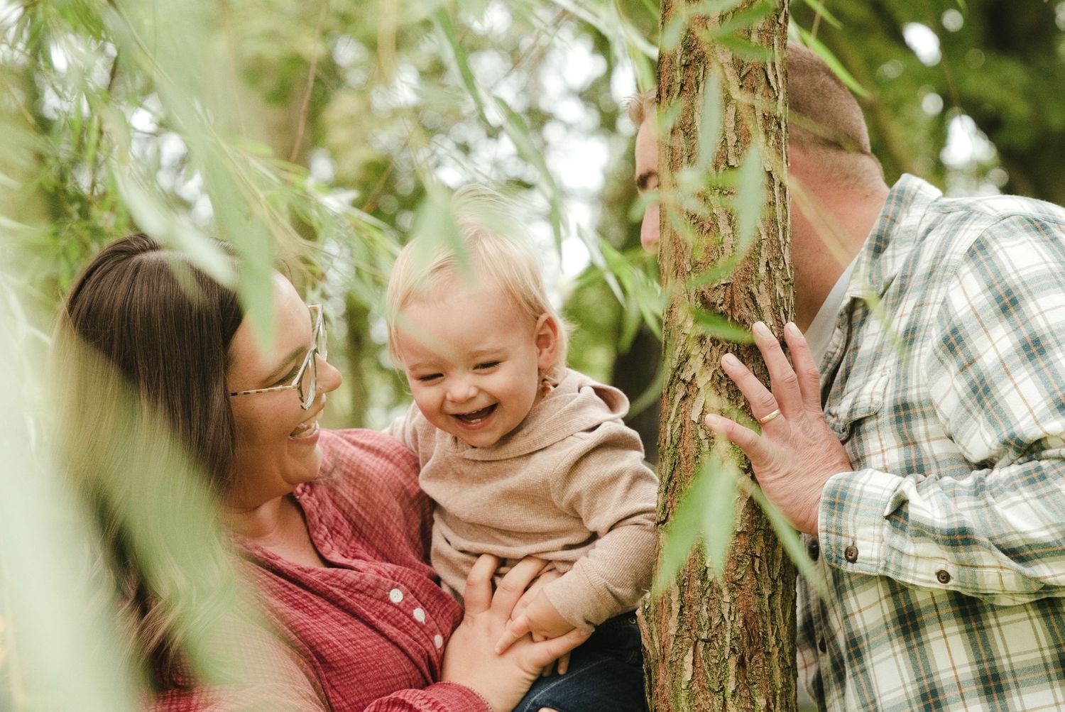 Smiling baby held by a woman in glasses and a man in a plaid shirt beside a tree trunk with green leaves in a sunny outdoor family portrait.