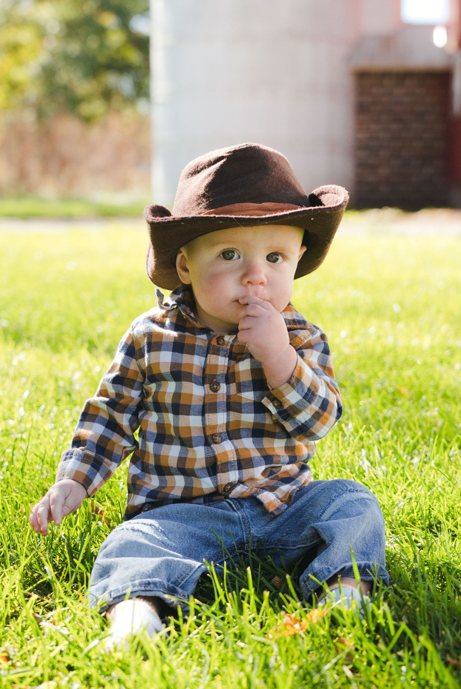 Baby in a brown cowboy hat and plaid flannel shirt sits on green grass with a hand at his mouth, a brick building behind him.