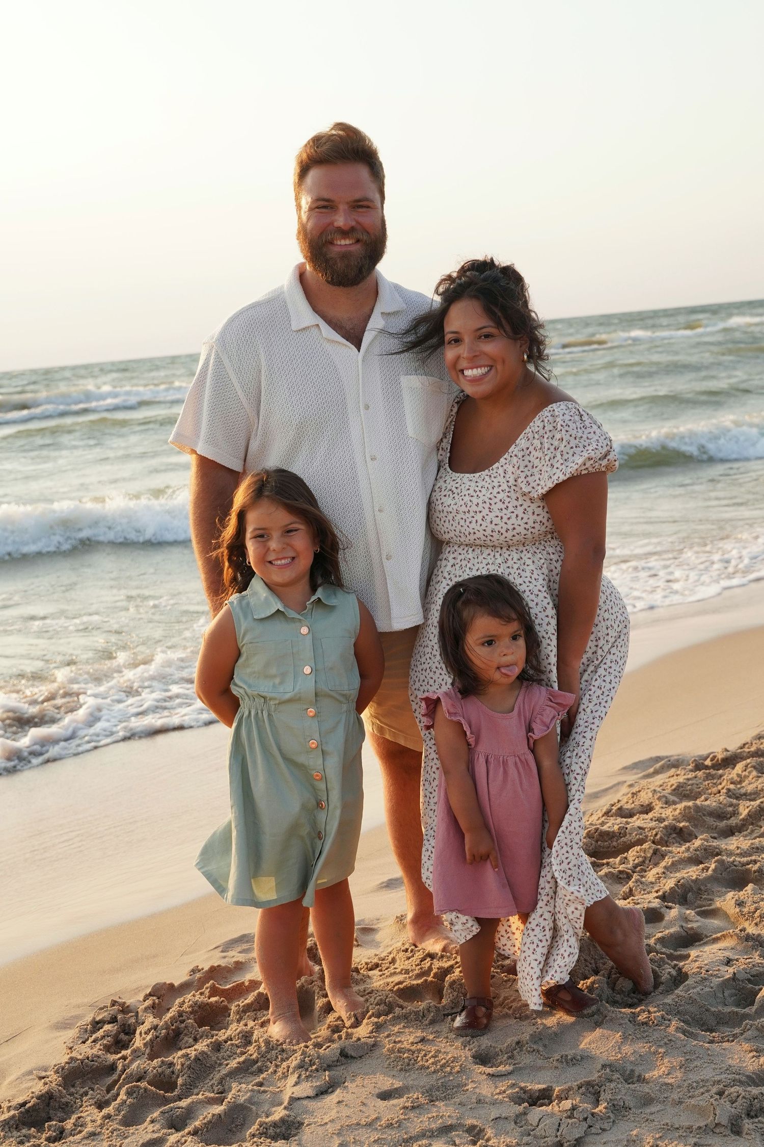 Family of four smiling together on a sandy beach at sunset, parents with two young daughters in summer dresses.