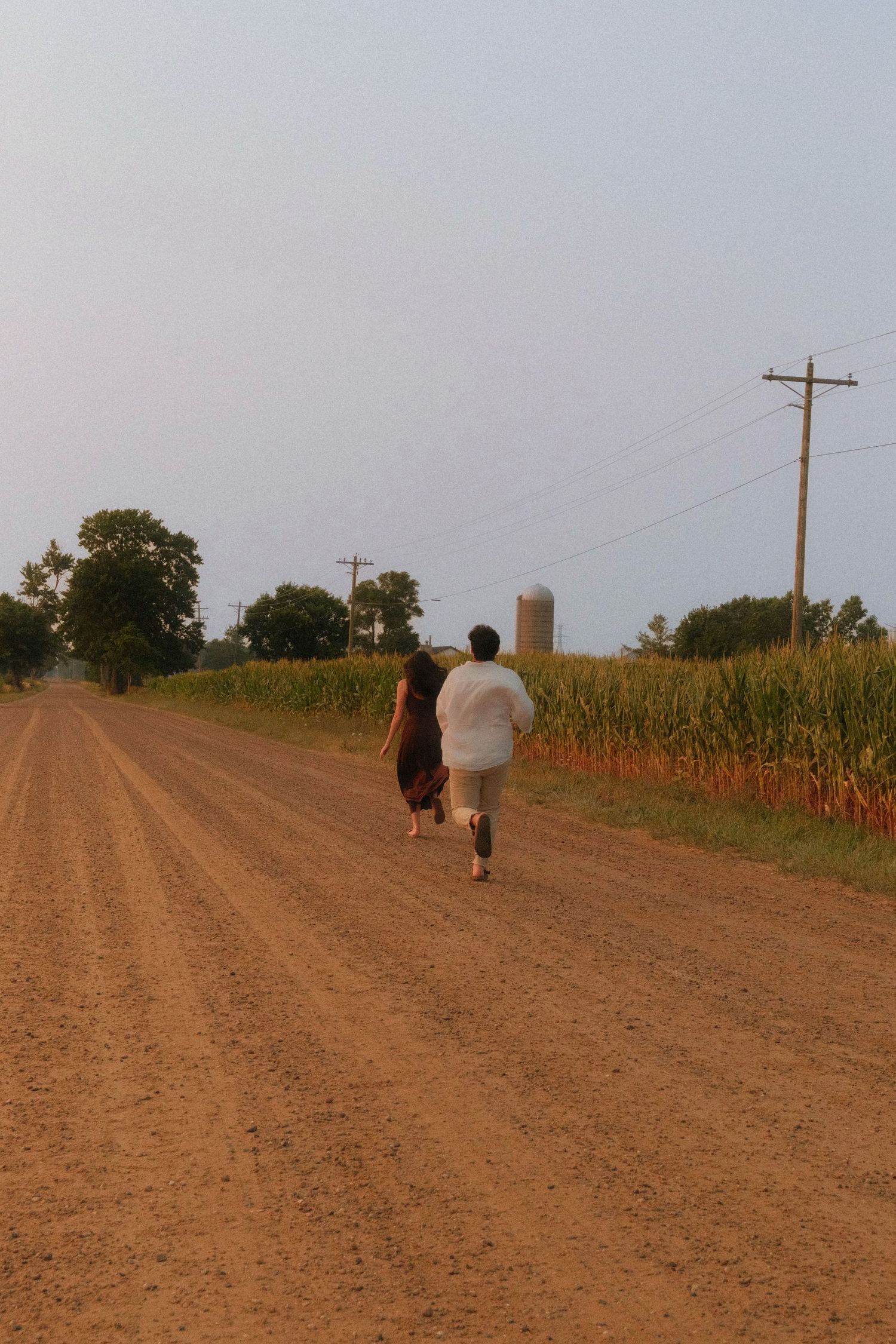 A couple walks hand in hand down a rural dirt road flanked by cornfields and utility poles at dusk.