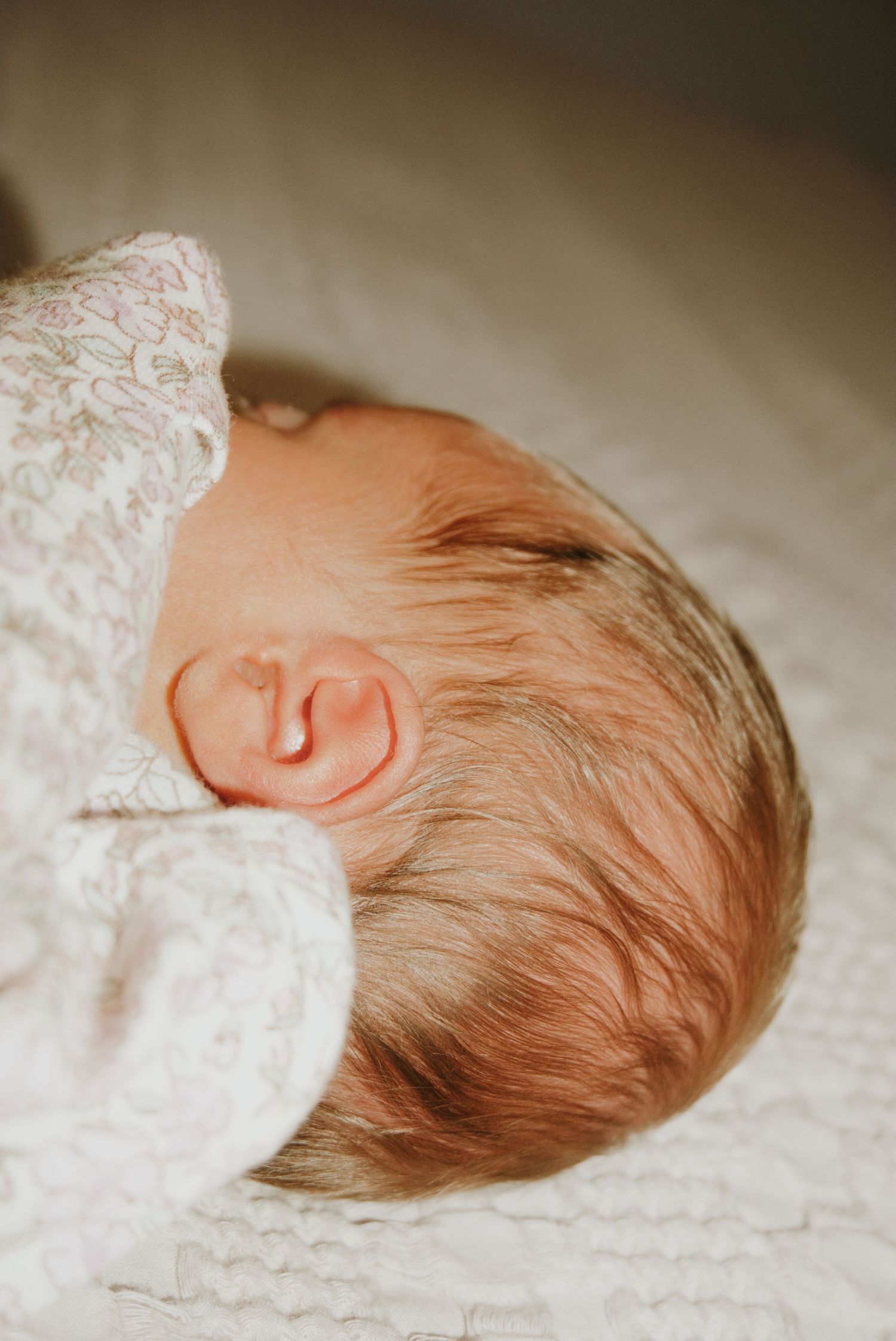 Close-up of a sleeping newborn baby with fine hair wearing a white lace outfit on a soft white blanket.