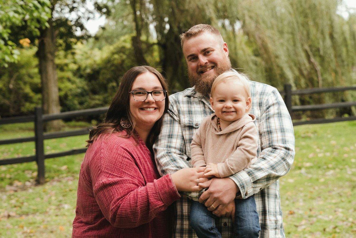 Smiling woman in a red sweater and bearded man in a plaid shirt hold a happy toddler outdoors near a wooden fence and trees.