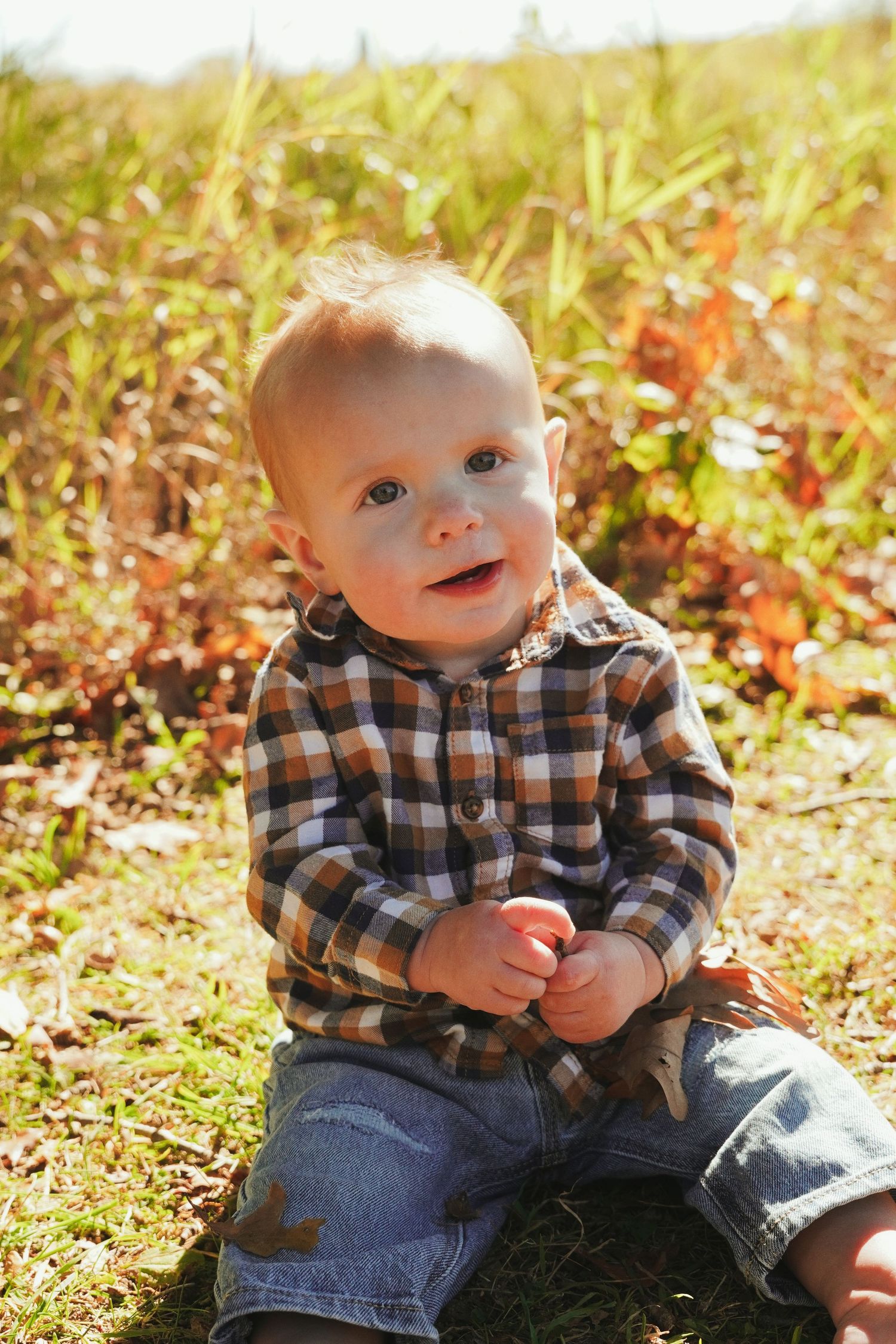 Baby boy in a plaid flannel shirt and jeans sits on grass, smiling and holding his hands together in a golden autumn field.