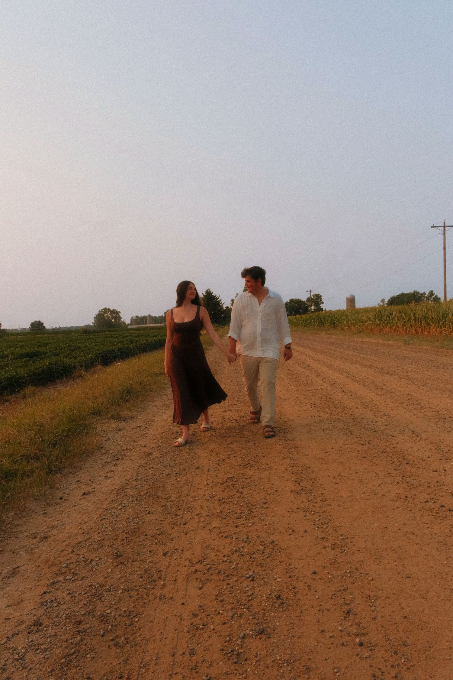 Couple holding hands walking along a scenic dirt road through farmland at dusk.