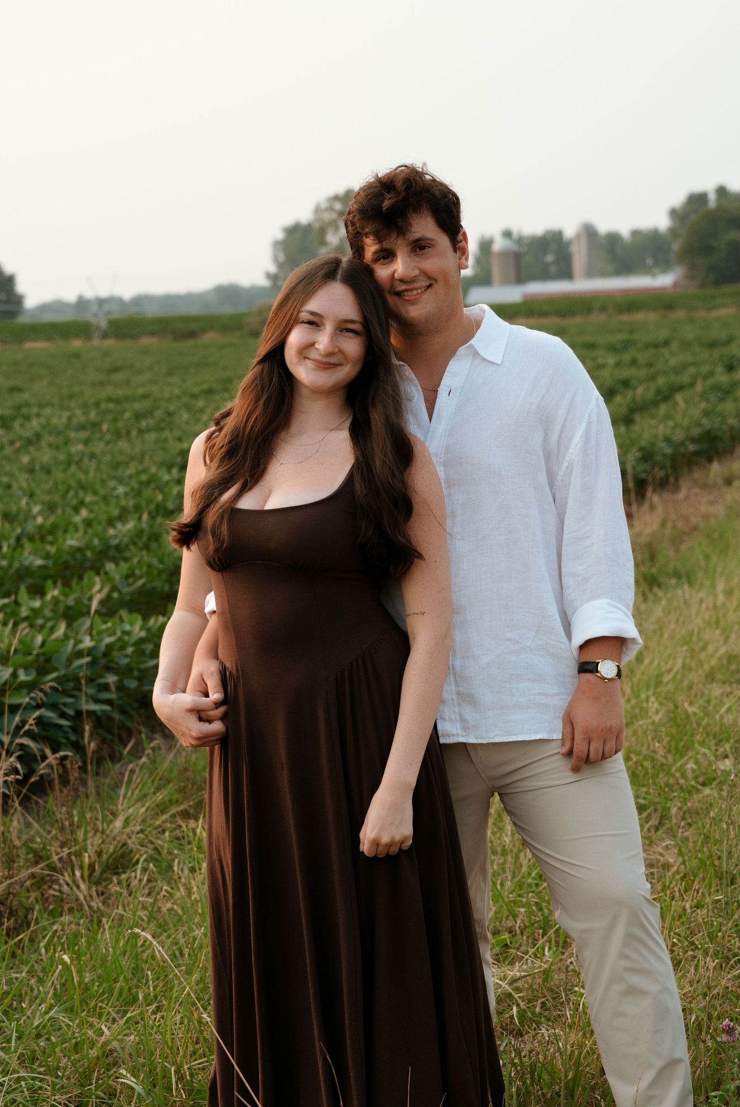 Smiling couple posing together in a green farm field, woman in brown maxi dress and man in white shirt.