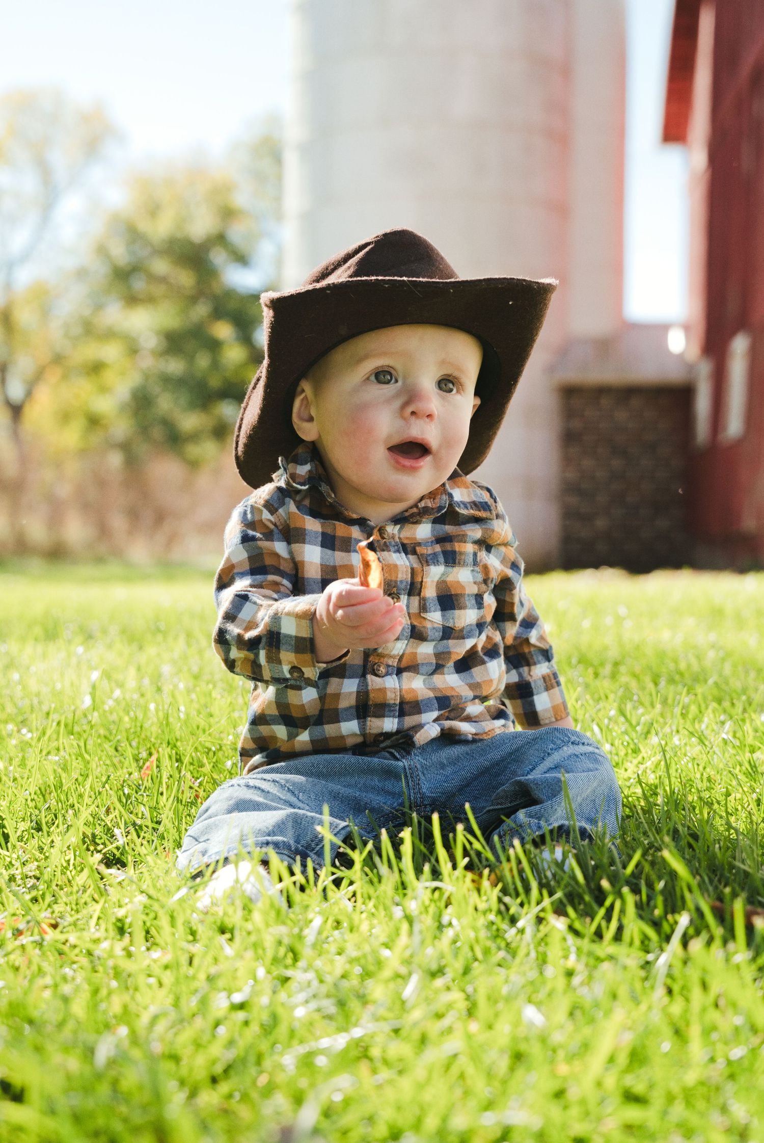 Toddler in a brown cowboy hat and plaid shirt sits on green grass holding a snack near a red barn and silo.