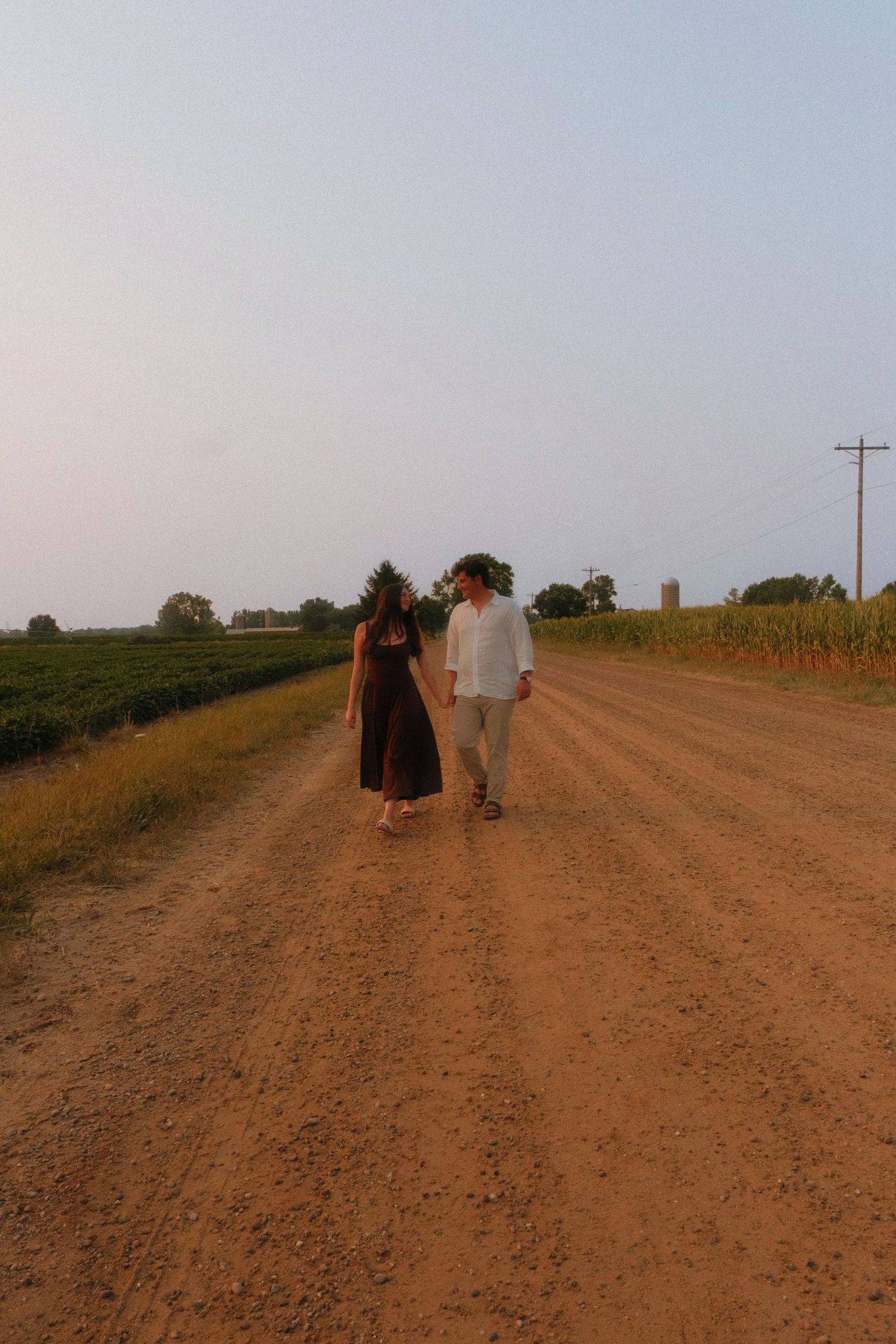 Couple walking hand in hand down a rural dirt road surrounded by farmland at dusk.
