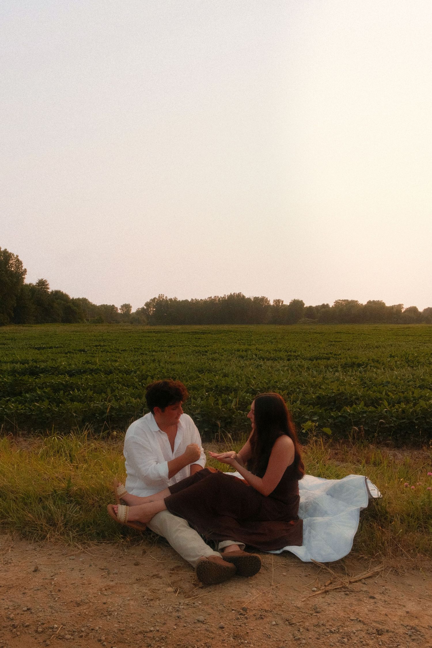 Couple sitting together on a dirt path beside a green farm field at dusk.
