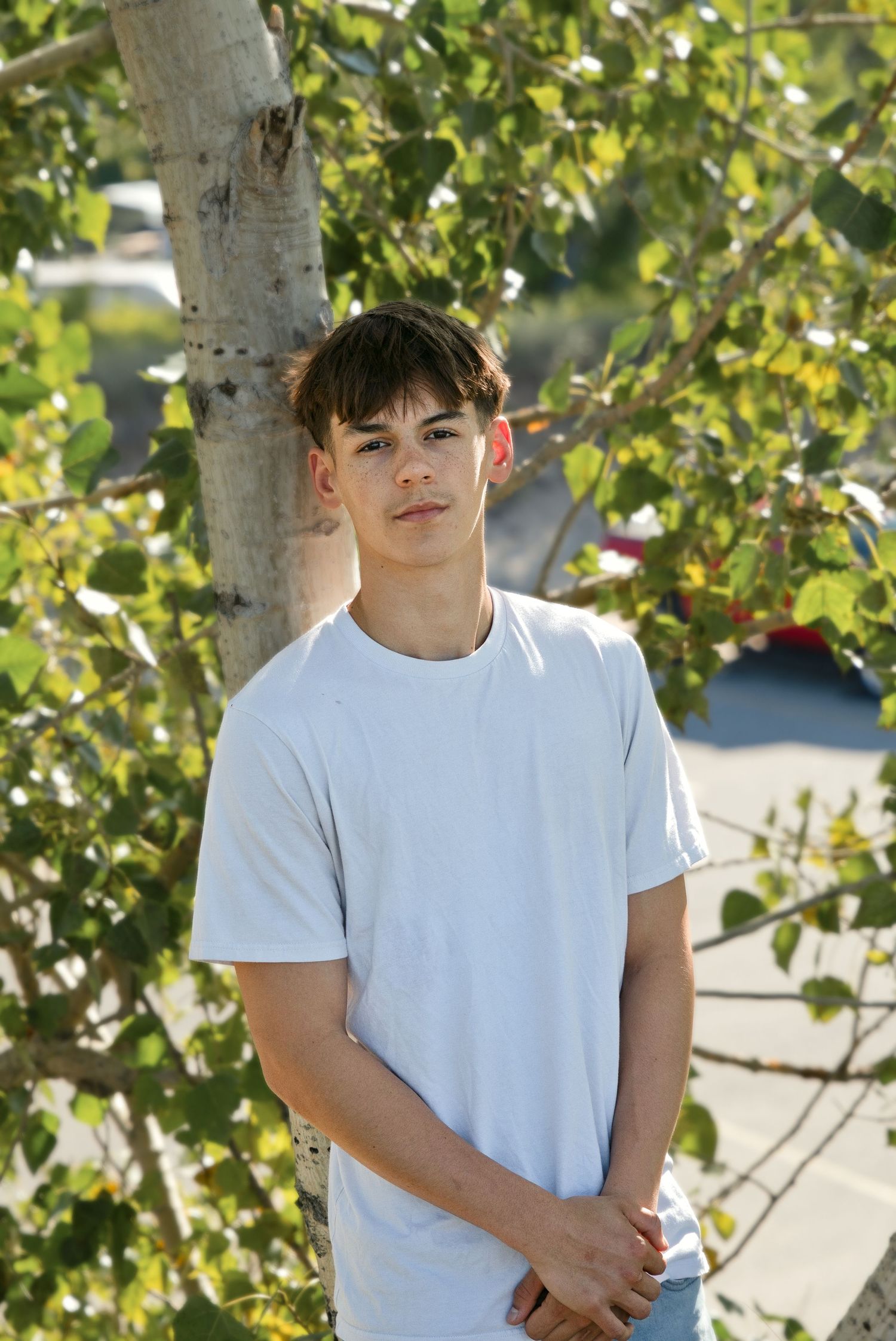Young teen boy in white t-shirt stands confidently by a tree with green leaves in a sunny outdoor setting.