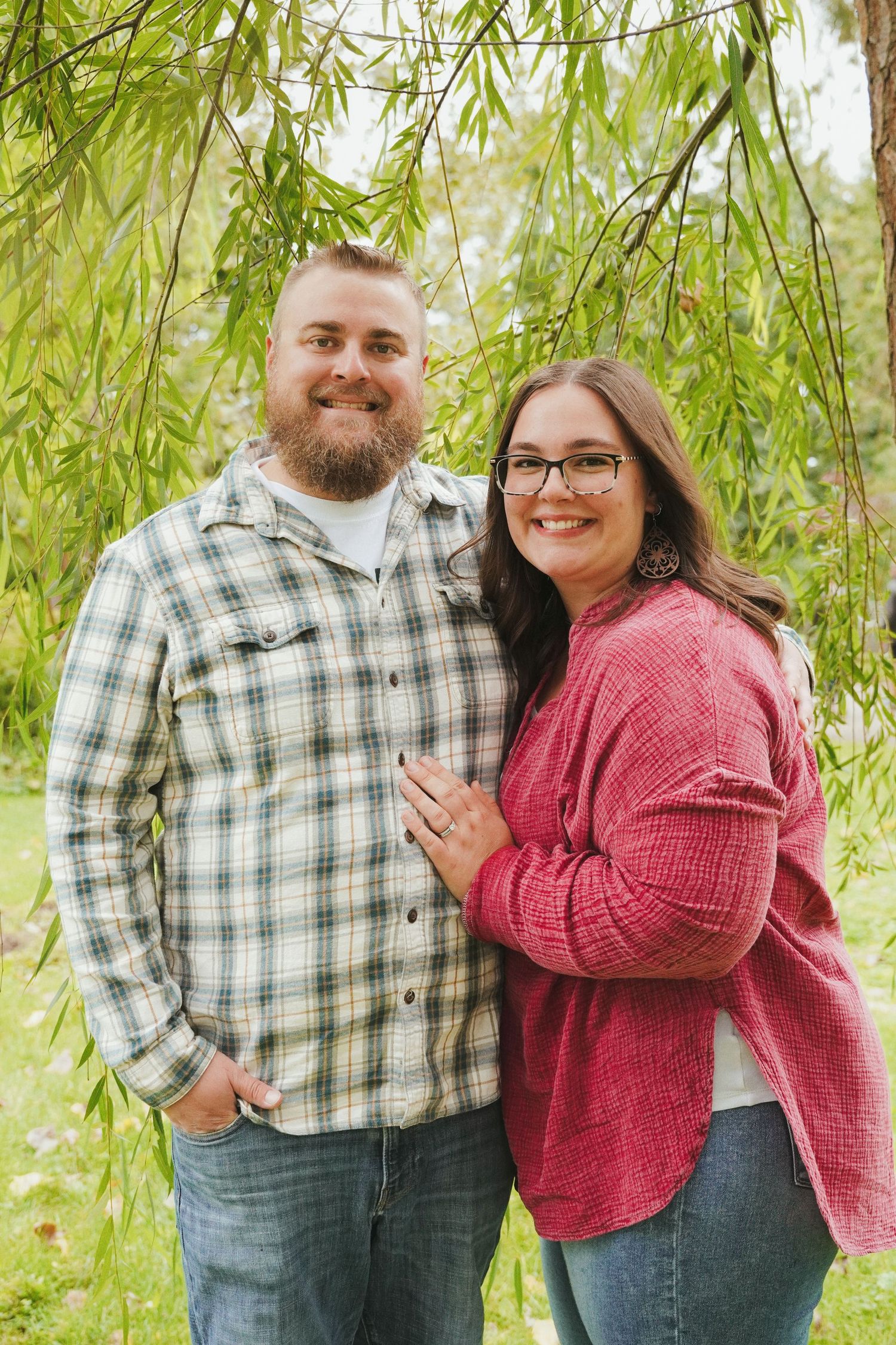 Smiling couple embracing outdoors beneath willow branches, he in a plaid shirt and jeans, she in a red cardigan and glasses.