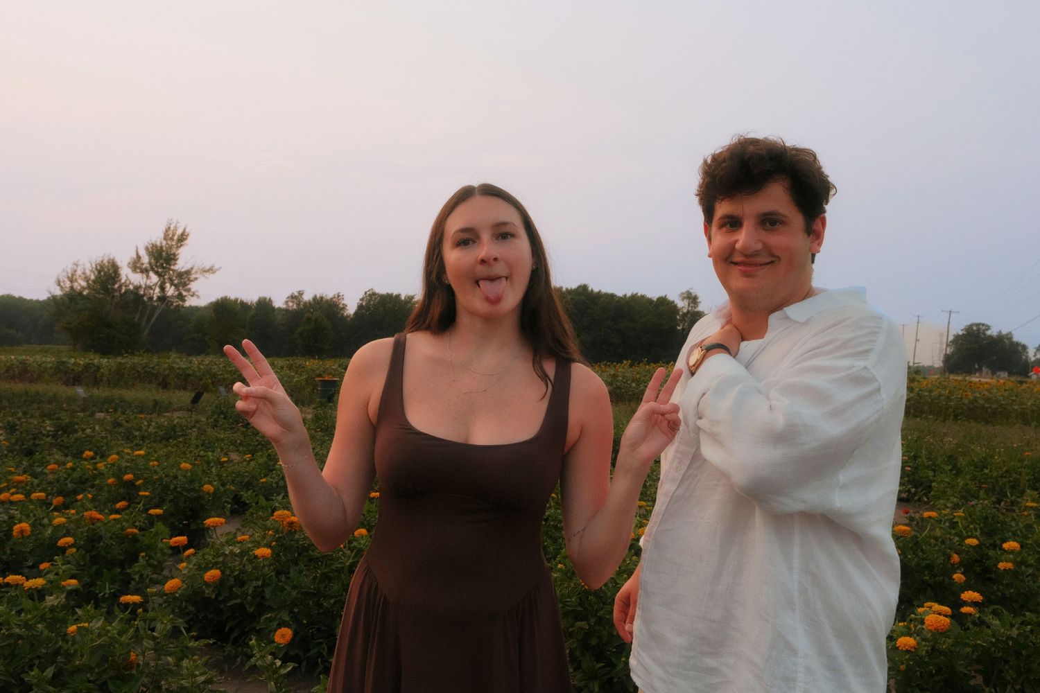 Two women smiling and making peace signs in a flower field at dusk.