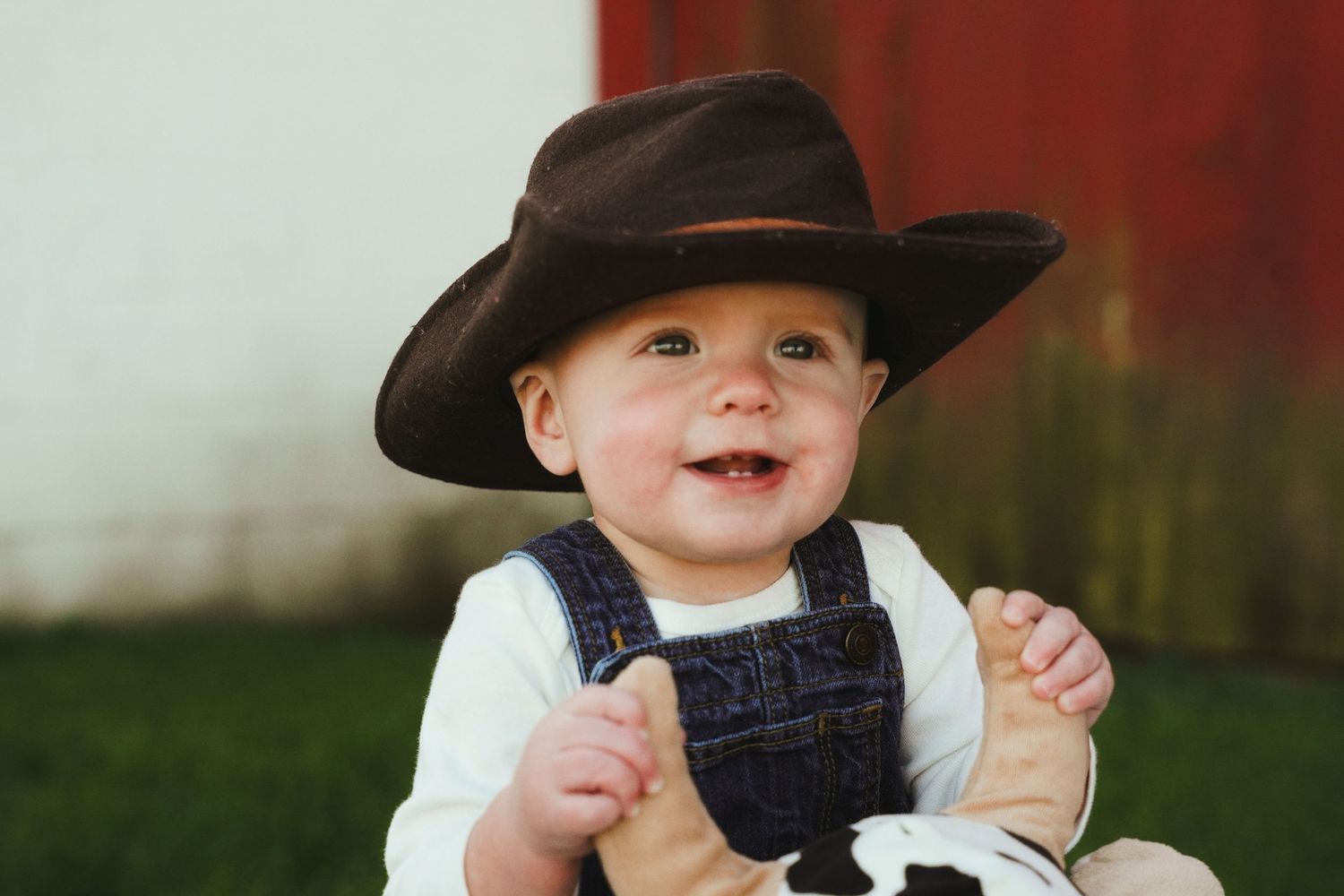 Smiling baby in a brown cowboy hat and denim overalls sits outdoors holding a foot, with a red barn wall and green grass behind.