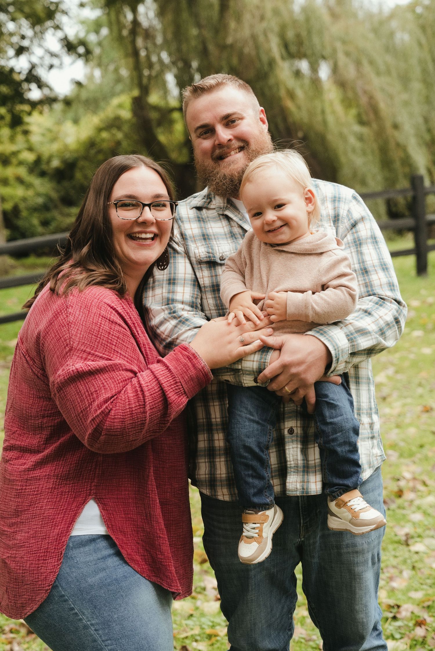 Smiling couple in plaid and red cardigan hold their laughing toddler in a family portrait outdoors beneath willow trees.