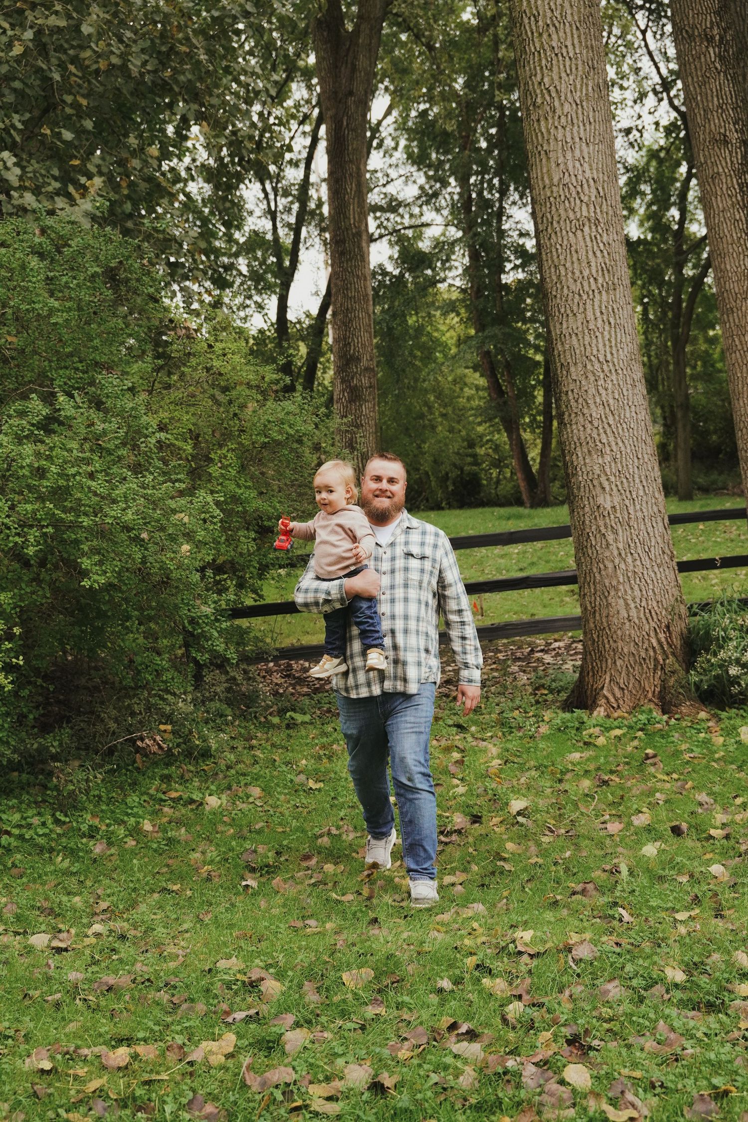 Bearded man in a plaid shirt and jeans carries a smiling toddler across a grassy park lined with tall trees and fallen autumn leaves.