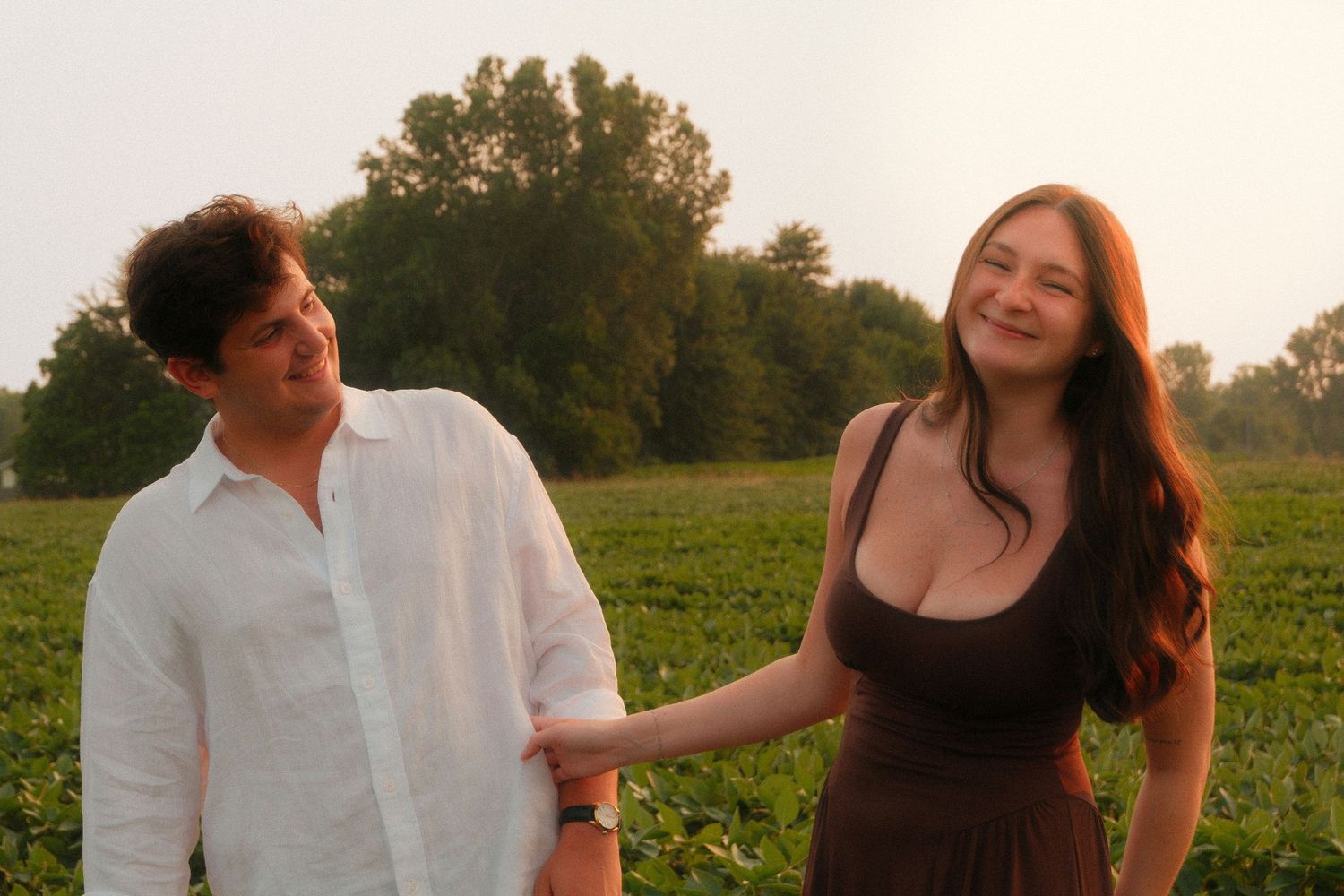 Couple holding hands smiling in a green field at golden hour, woman in brown dress, man in white shirt.