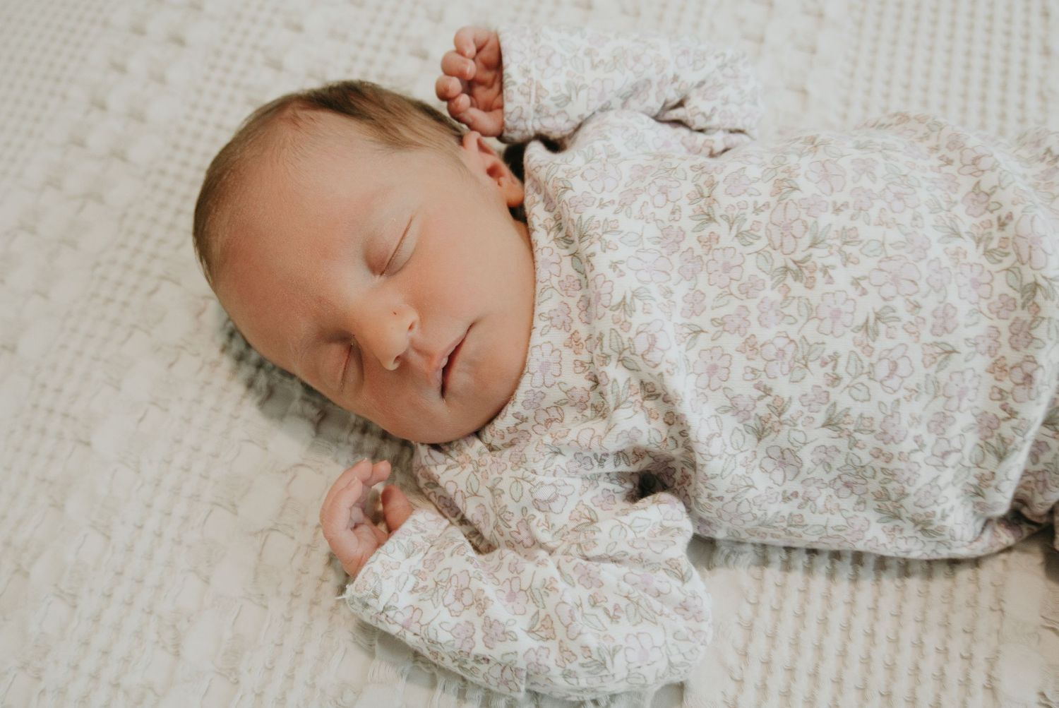 Peaceful newborn baby sleeping soundly in a floral onesie on a soft white textured blanket.