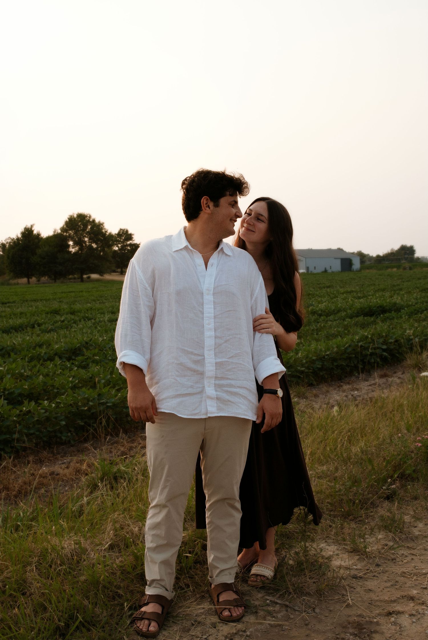 Couple walking together on a dirt path beside a green farm field at sunset.