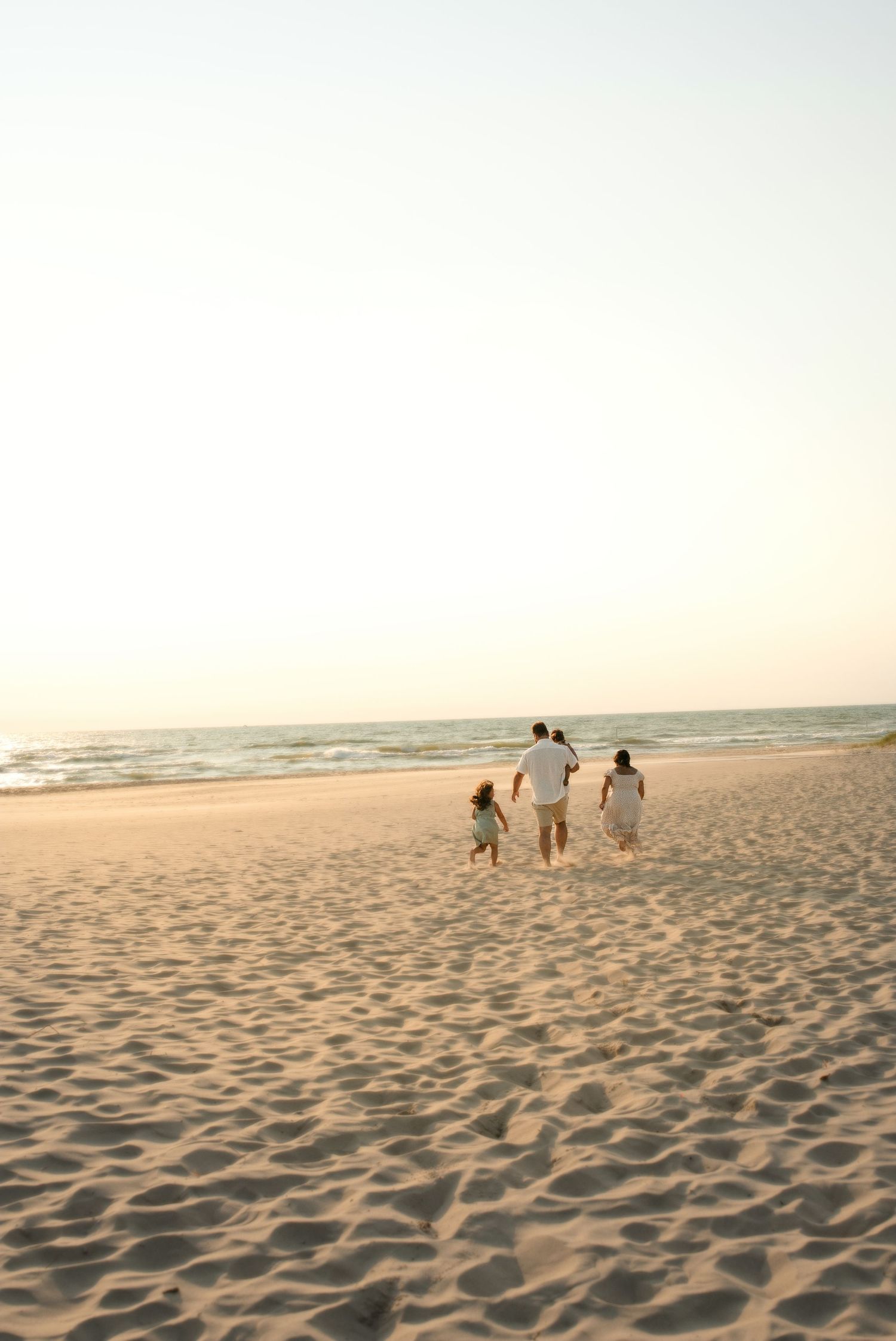 A family of four walks hand-in-hand toward the ocean on a wide sandy beach at sunset.