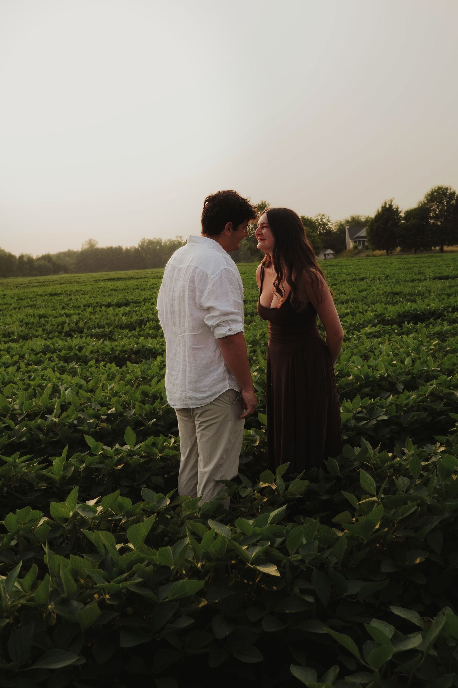 Romantic couple standing face to face smiling in a green crop field during a golden hour sunset.