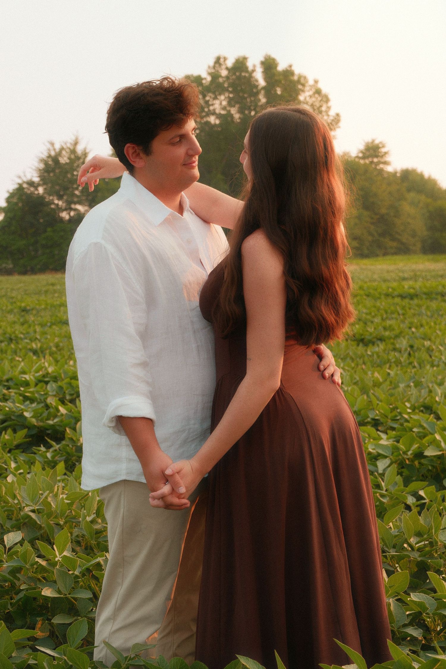 Couple holding hands and embracing in a lush green field at golden hour, woman in brown dress, man in white shirt.