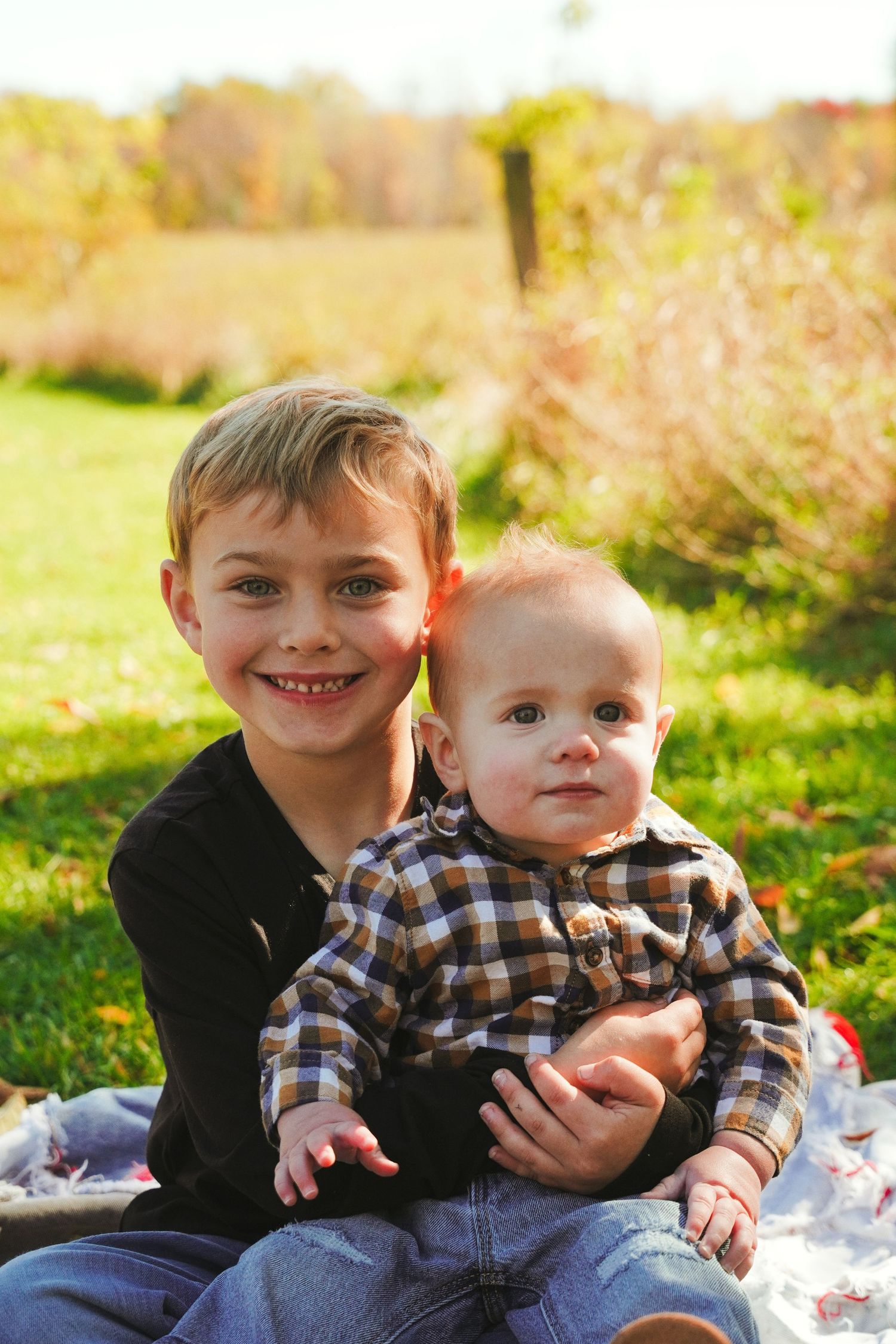 Close-up of a smiling boy holding a baby in a plaid shirt outdoors on a quilt with colorful fall foliage in the background.