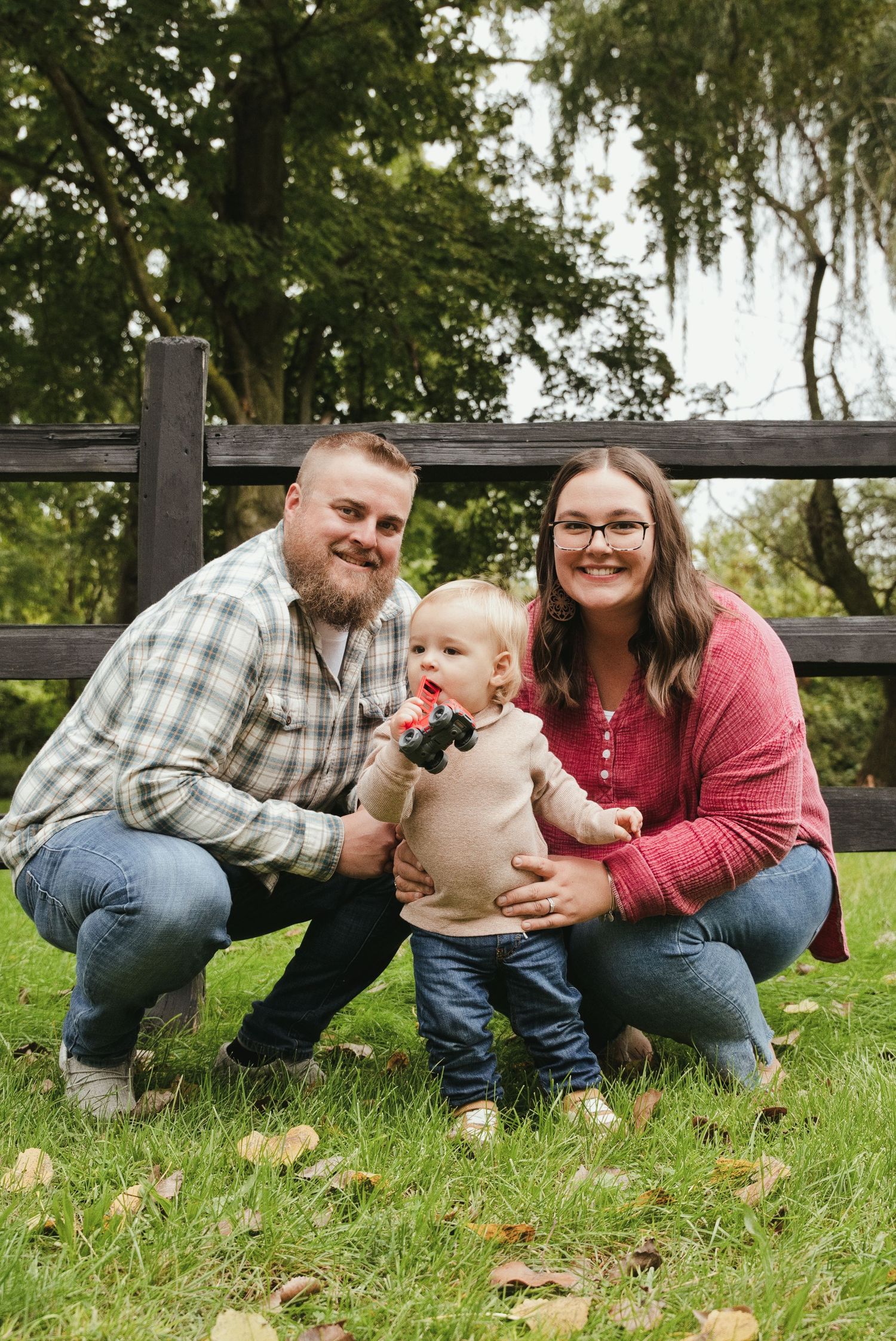 Family of three crouches together on the grass in front of a wooden fence, with the toddler holding a small toy.