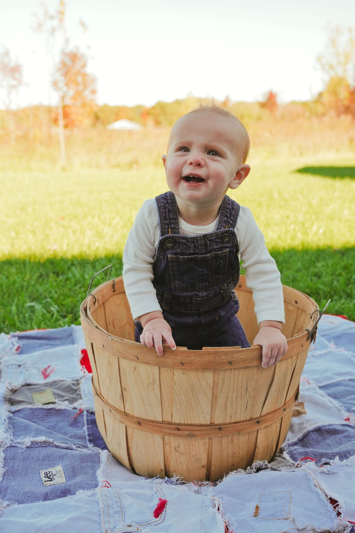 Smiling baby in denim overalls standing in a wooden bushel basket on a patchwork quilt in a sunny autumn field.