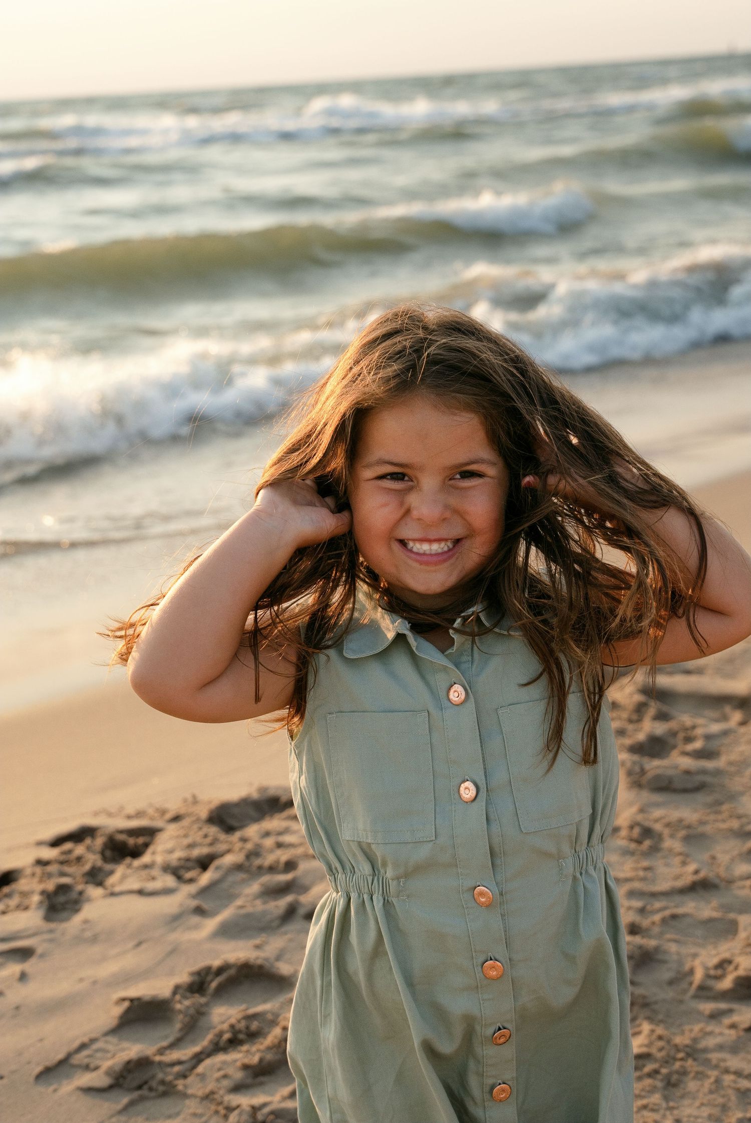 A smiling young girl in a green dress plays with her hair on a sandy beach with ocean waves behind her.