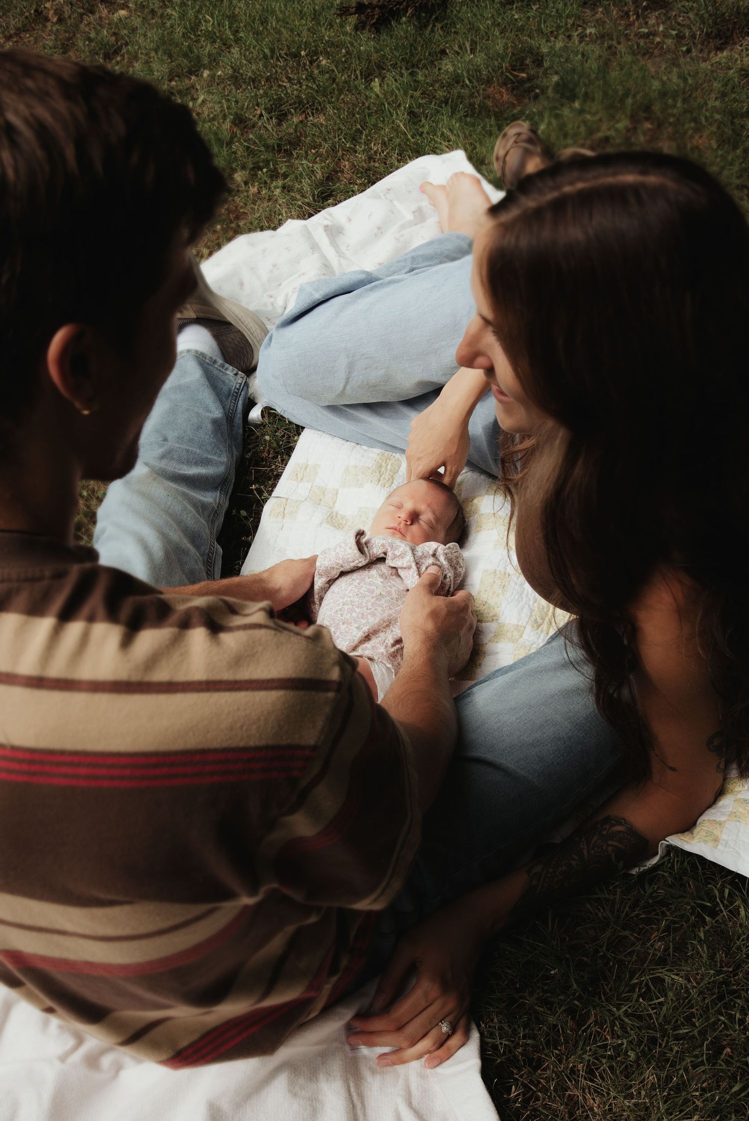 Two parents sitting on a blanket outdoors, lovingly gazing at their newborn baby in a sunny garden setting.