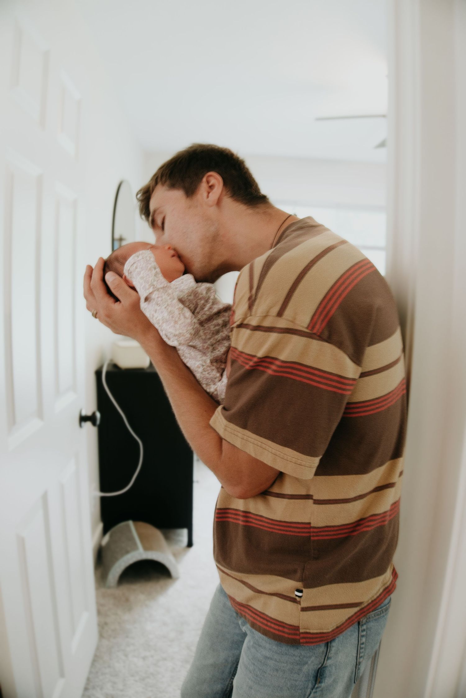 A young father in a striped shirt tenderly kisses his newborn baby while leaning against a white wall.