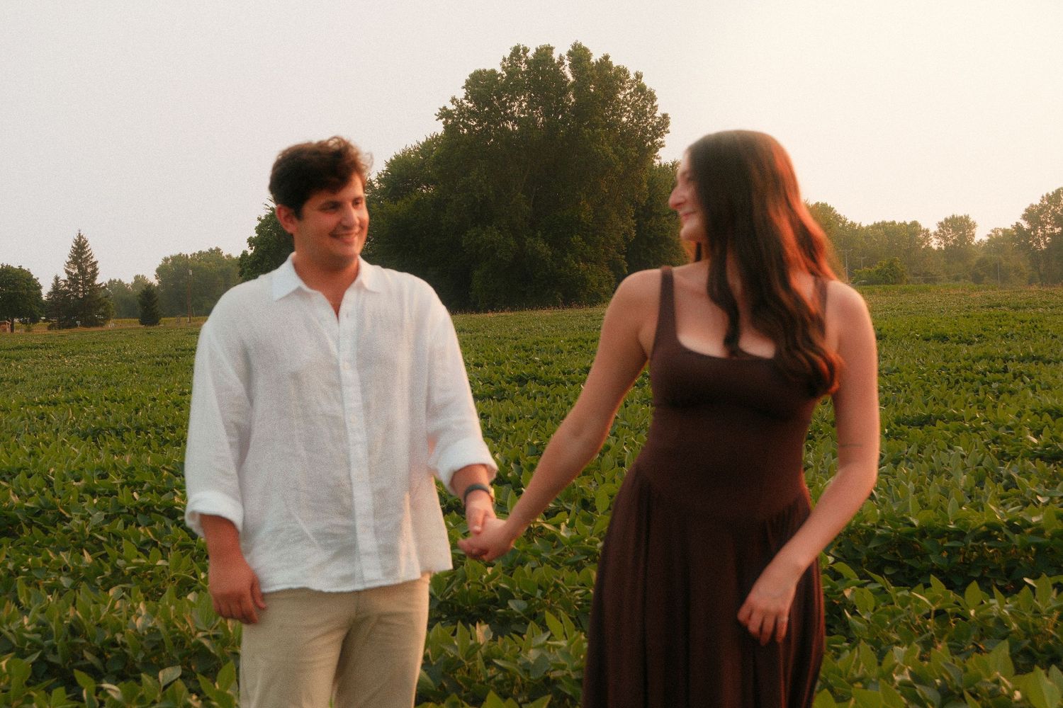 Couple holding hands in a lush green field, man in white linen shirt and woman in brown dress.