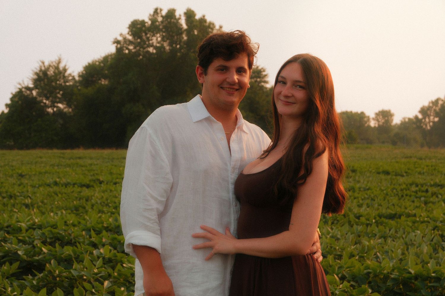 A couple smiles together in a lush green field at sunset, man in white shirt and woman in brown dress.