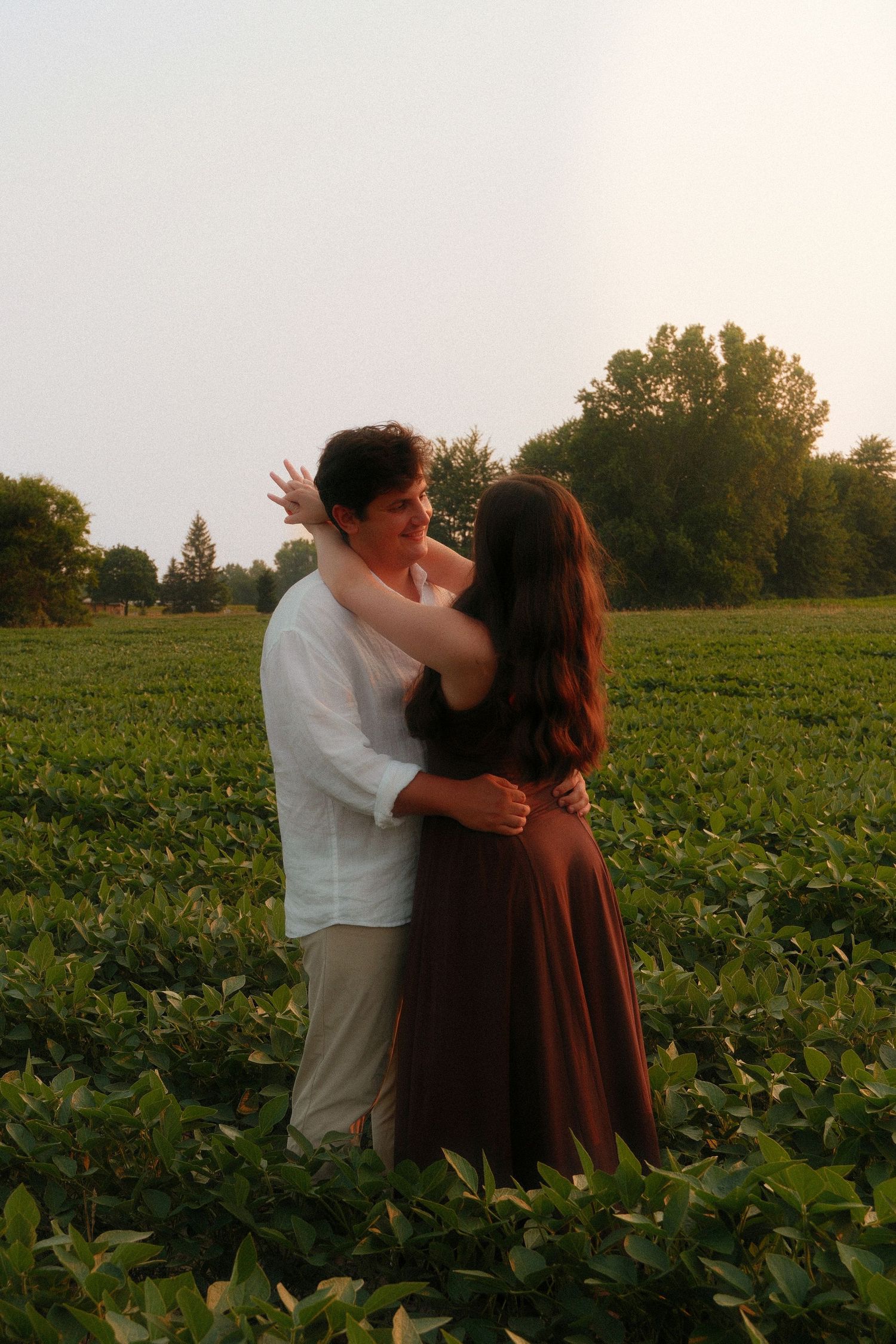 A couple embraces romantically in a lush green field at sunset, the woman touching the mans face tenderly.